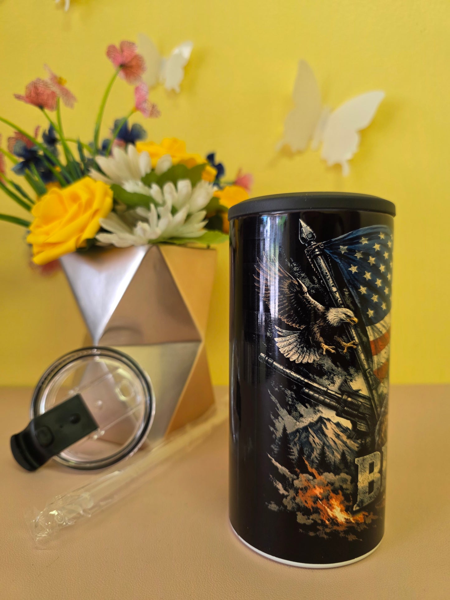 Black insulated stainless-steel tumbler with eagle and American flag artwork, shown with lid, straw, and floral background