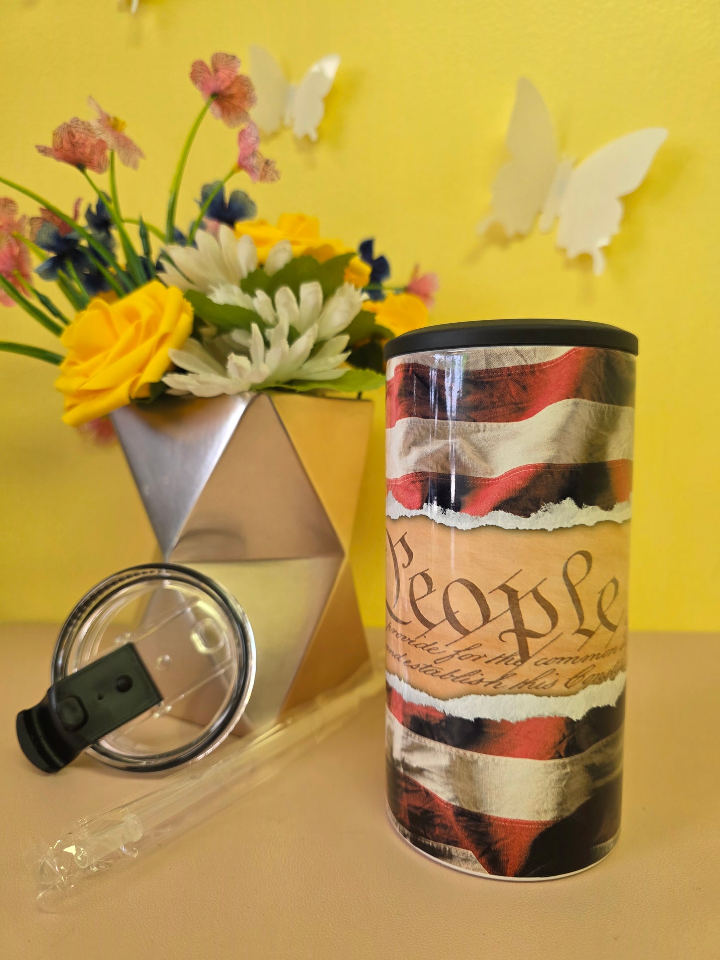 Insulated 4‑in‑1 stainless steel tumbler with U.S. Constitution text and American flag design, shown with lid, straw, and floral arrangement