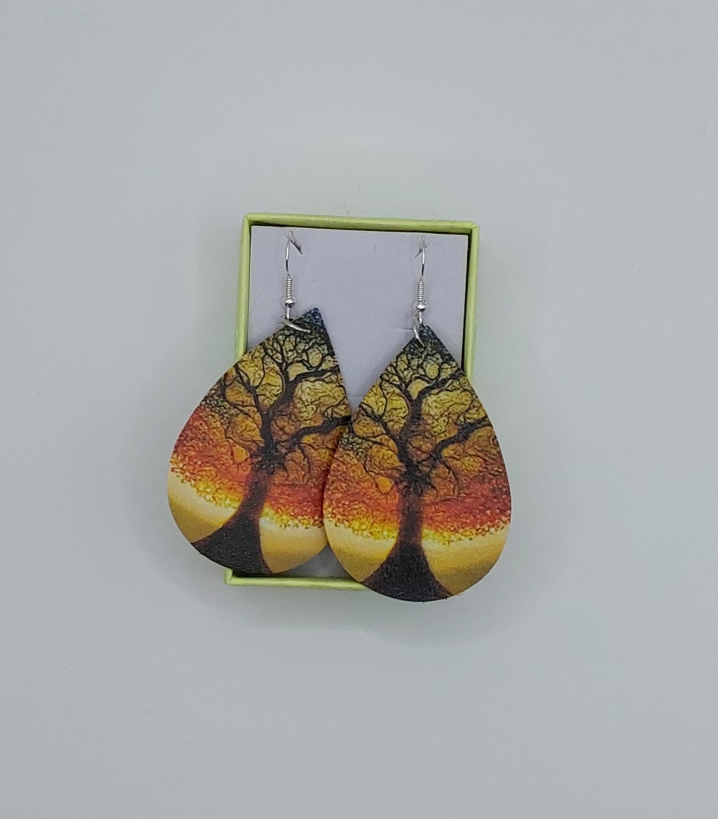 A pair of teardrop-shaped earrings with a autumnal tree pattern displayed in a small green box.