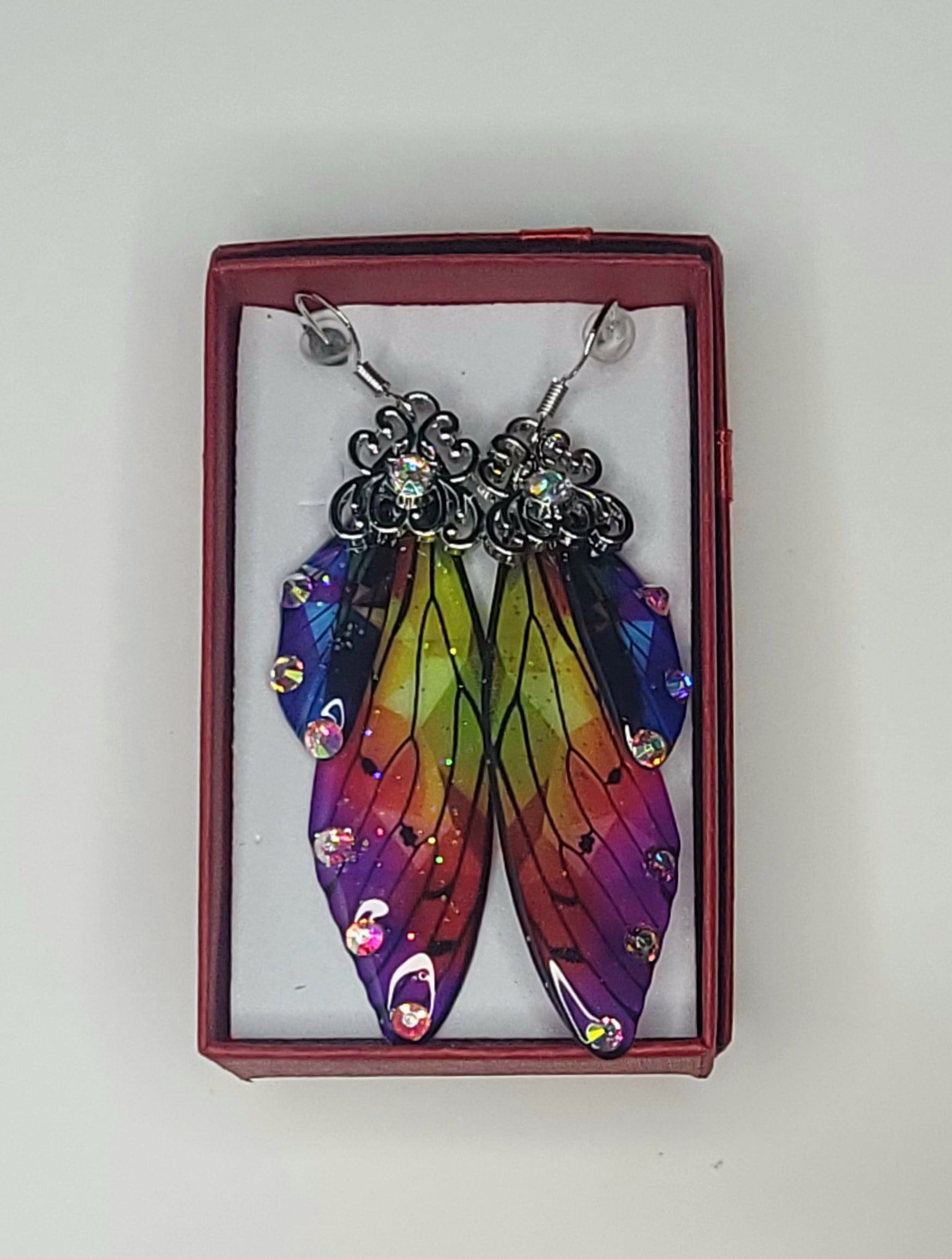 A collection of colorful butterfly wing earrings displayed in small boxes, arranged in a grid pattern with a candle lantern in the background.