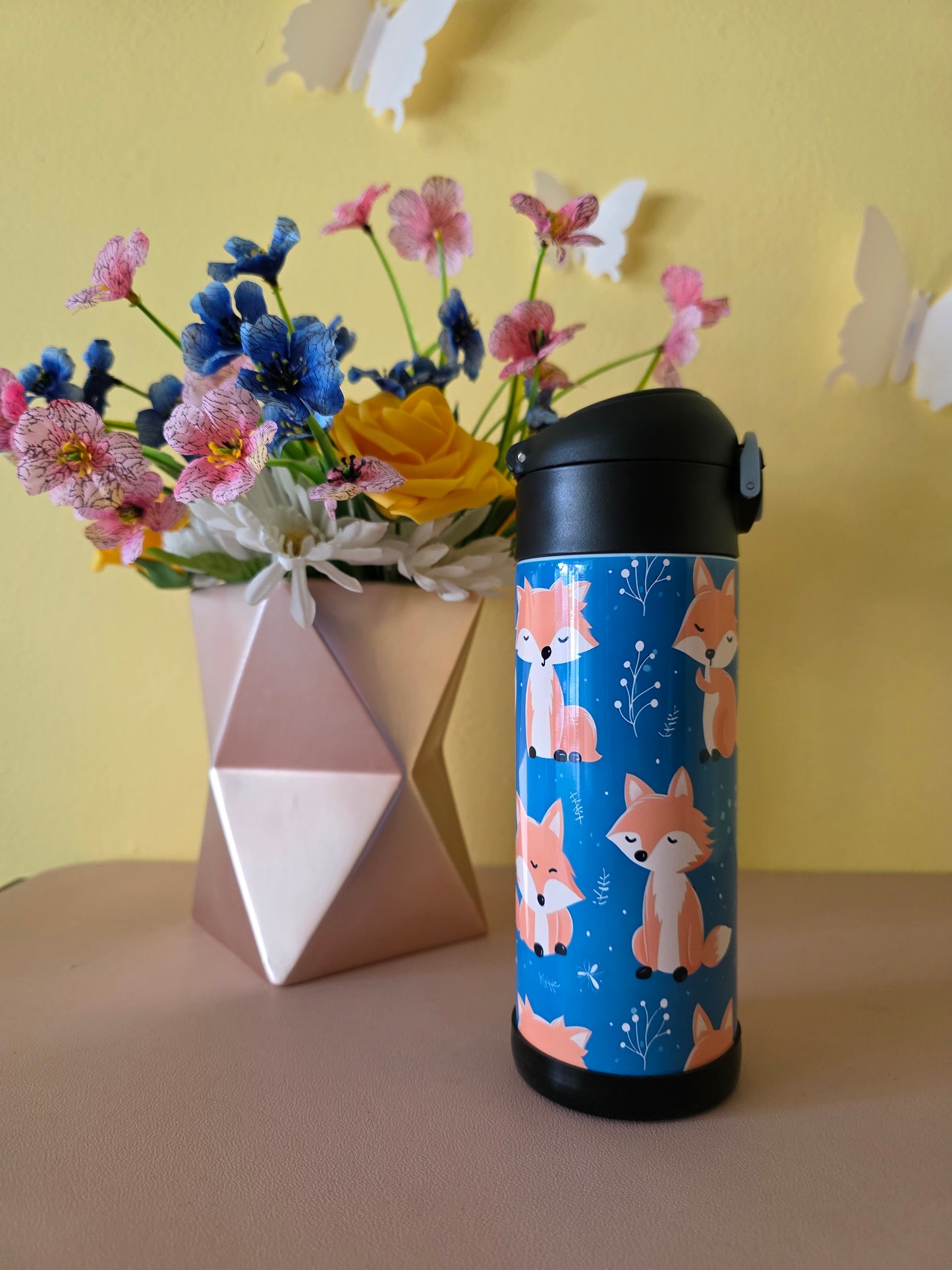 Thermos bottle with fox design next to a bouquet of flowers on a yellow surface