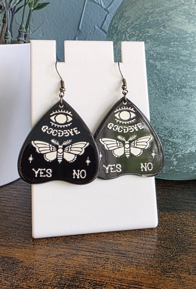 Black and white Quija planchette earrings with butterfly designs and 'Goodbye' text on a white stand