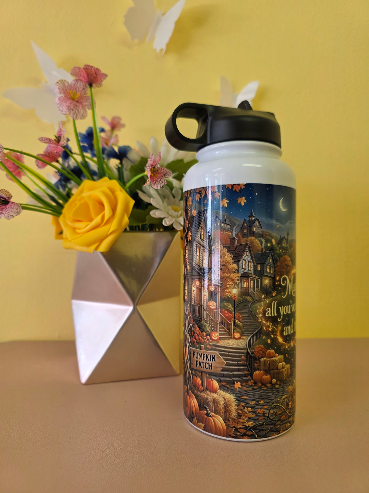 A white insulated water bottle with a black lid stands on a beige surface against a yellow wall. The bottle features a printed autumn village scene with pumpkins, houses, stars, and the text 'Merry all your days with happiness' and 'Pumpkin Patch'.