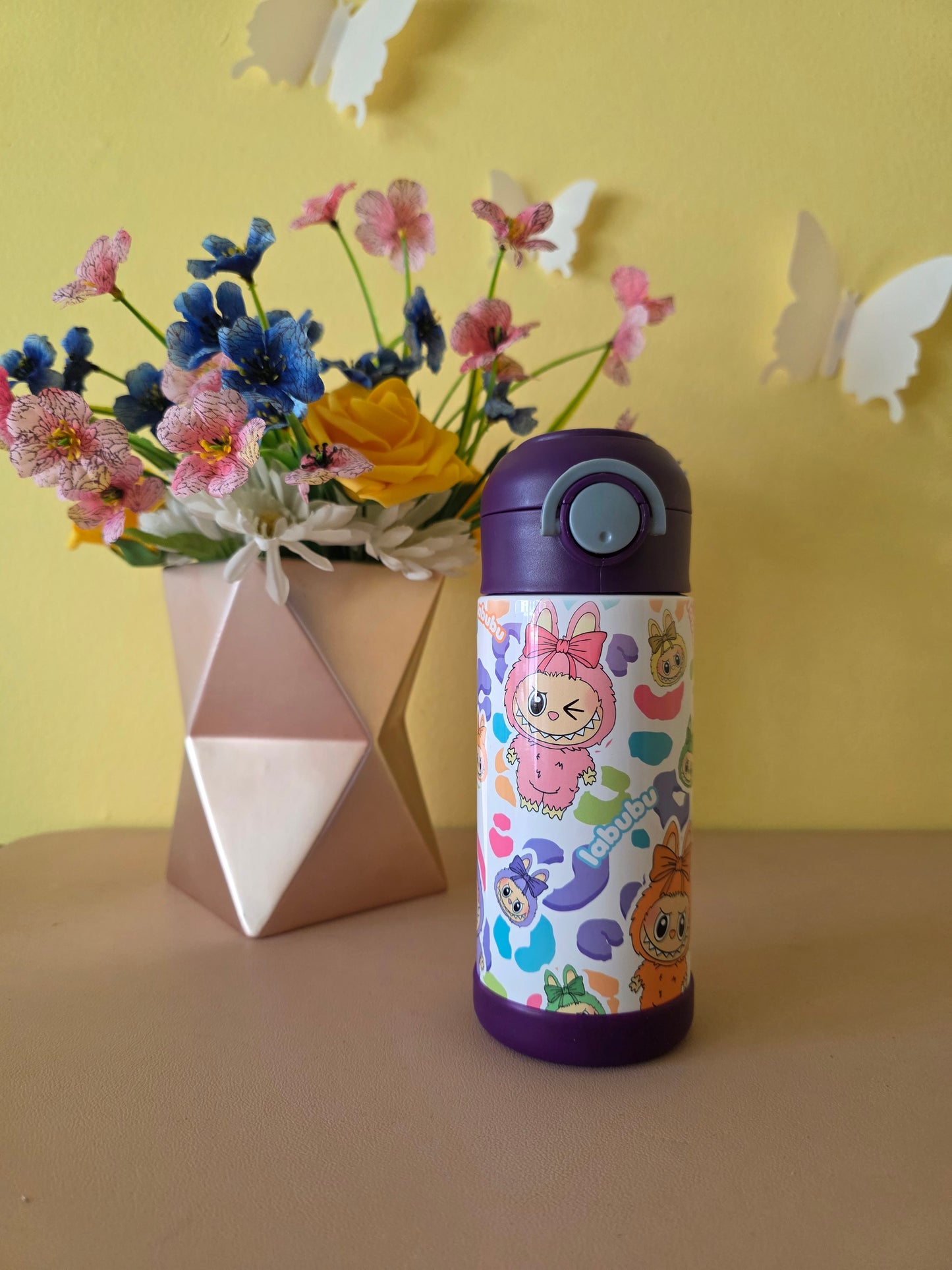 Colorful thermos with cartoon characters next to a bouquet of flowers on a yellow surface.