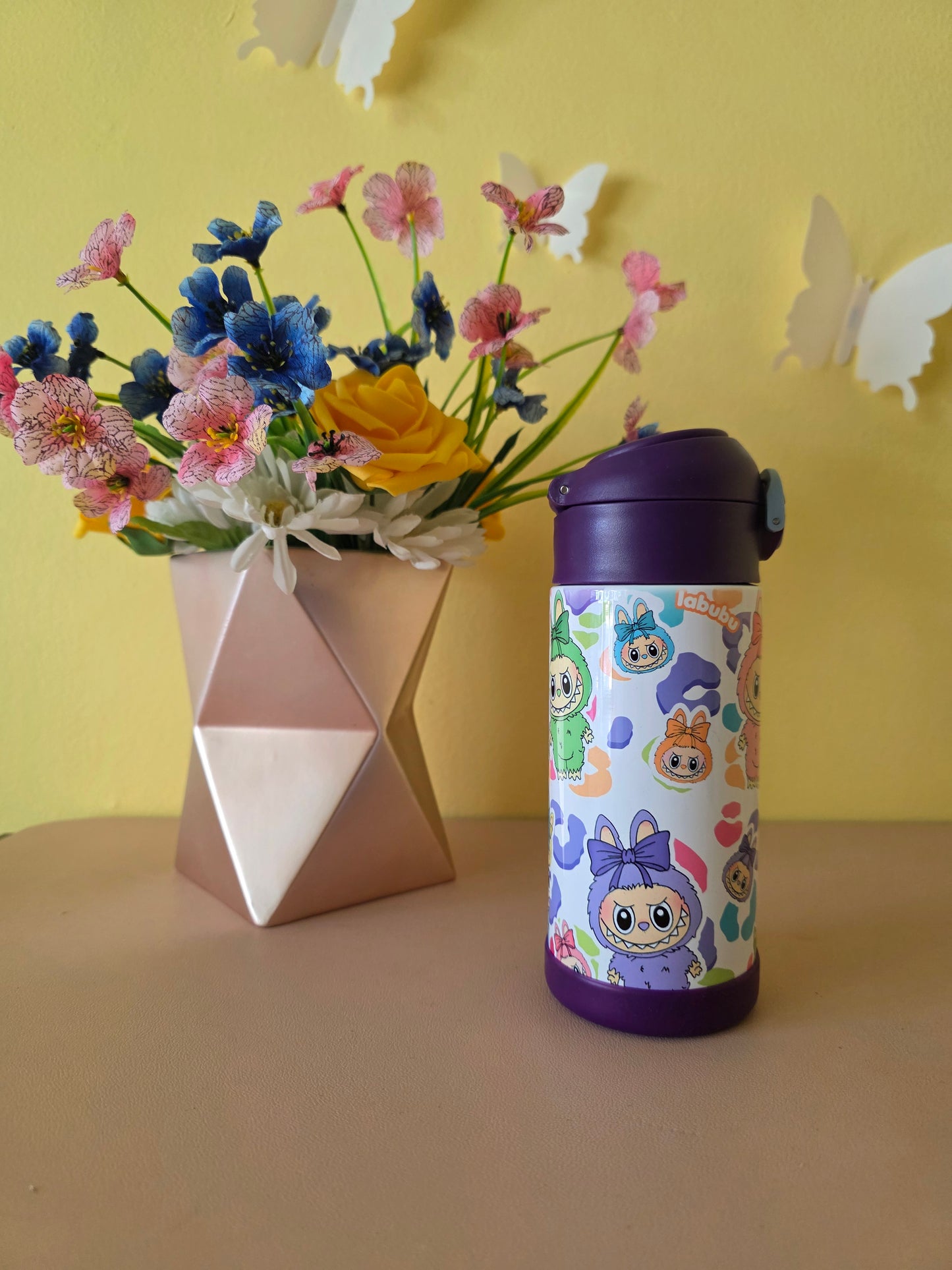 Colorful water bottle with cartoon design on a yellow surface with flowers and a box.