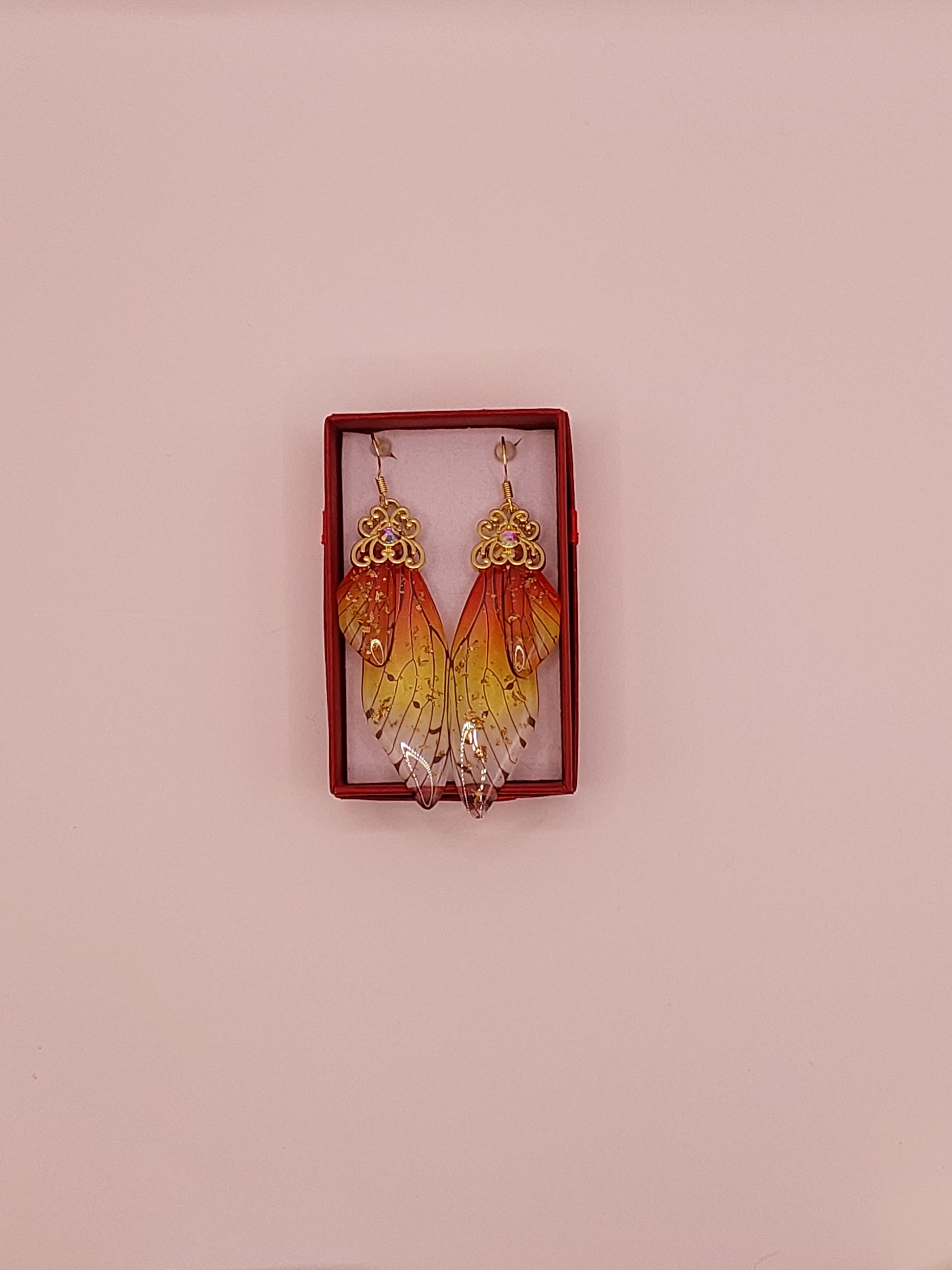 A pair of orange and gold butterfly wing earrings displayed in a red box on a pink background.