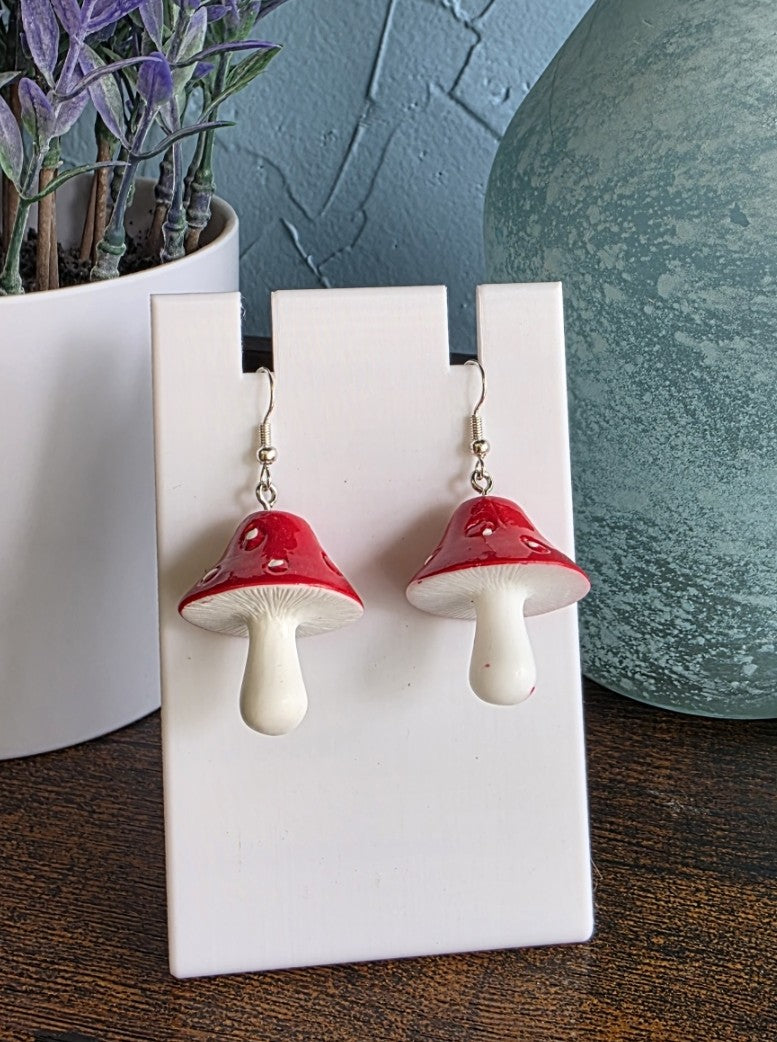 Red and white mushroom-shaped earrings on a white stand with a plant and vase in the background.