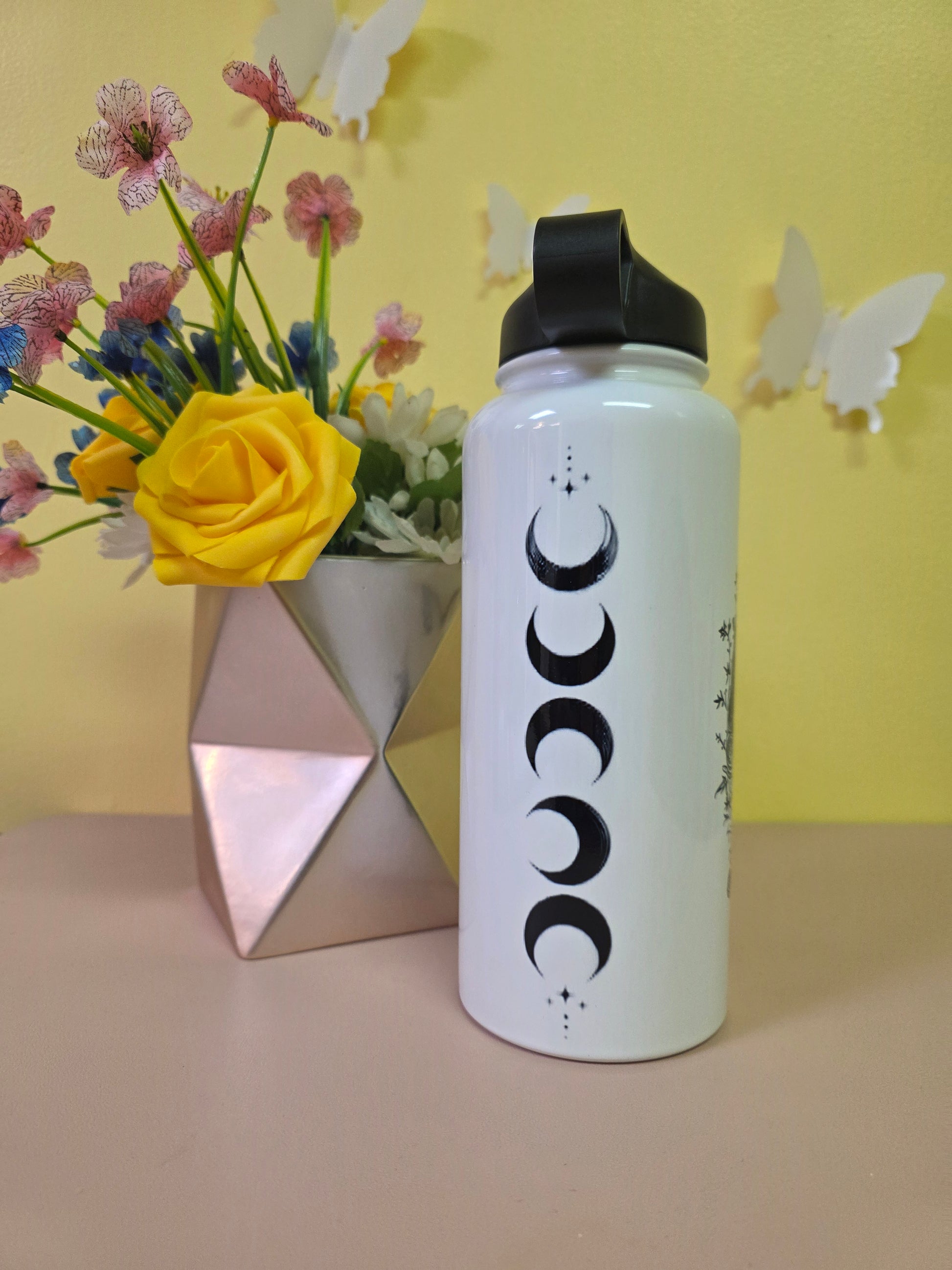 White water bottle with black crescent moon symbols on a yellow background with flowers and butterfly decorations.