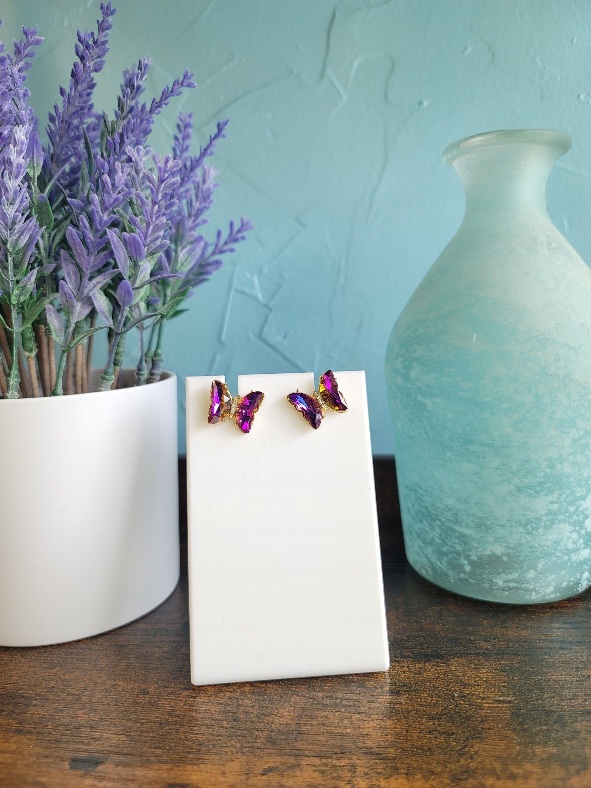 A pair of blue butterfly earrings with purple beads displayed on a white stand, with a lavender plant and a blue vase in the background.