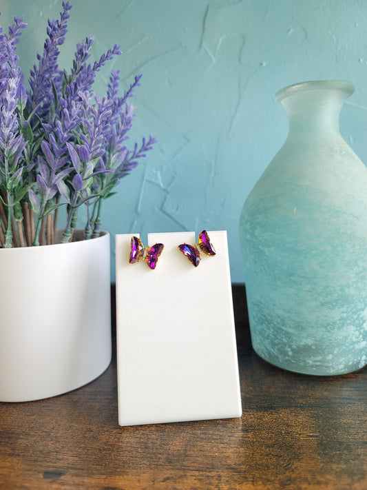 A pair of blue butterfly earrings with purple beads displayed on a white stand, with a lavender plant and a blue vase in the background.
