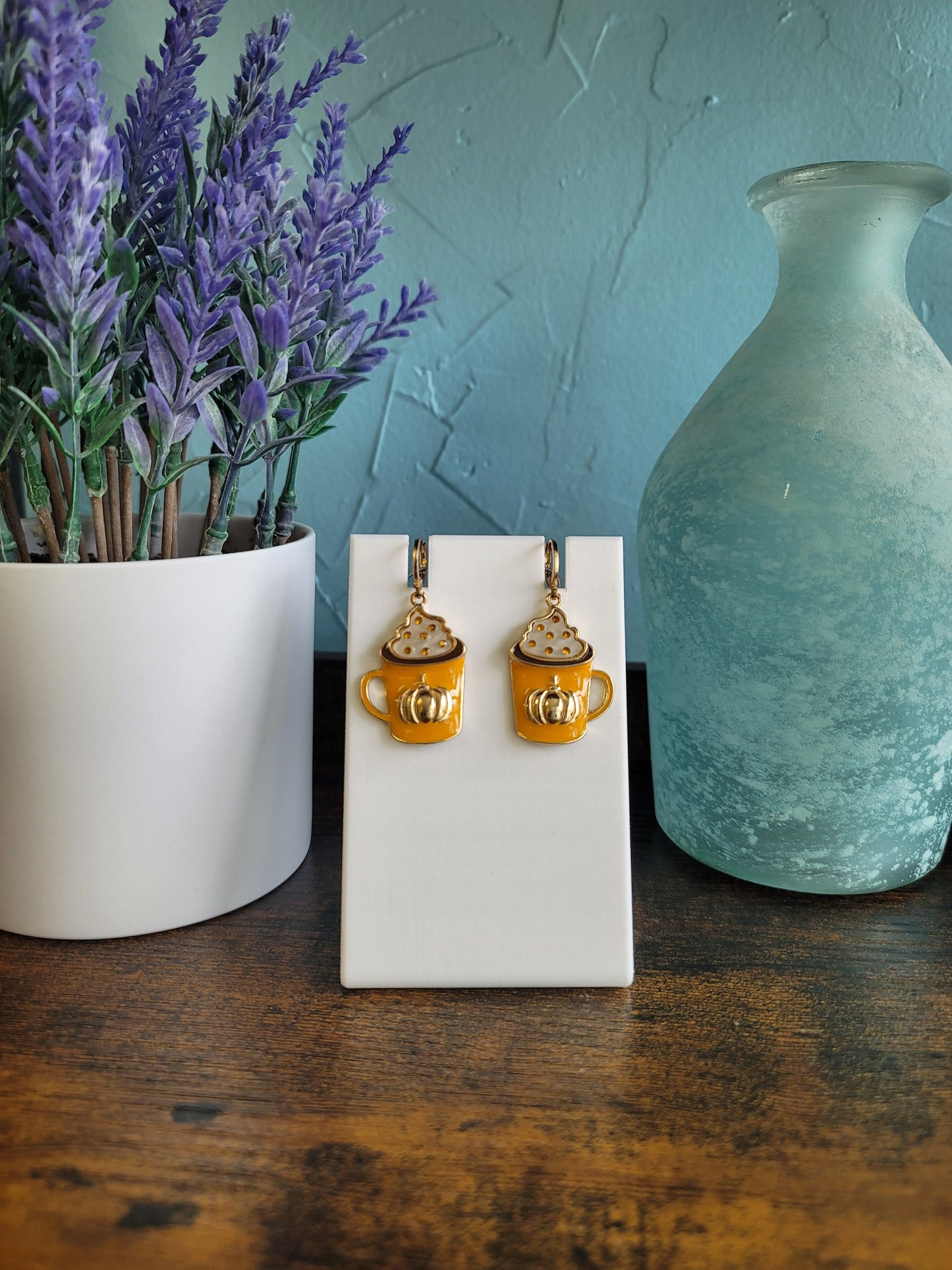 A pair of gold-colored earrings designed to resemble coffee beans, displayed on a white stand, with a lavender plant and a blue vase in the background.