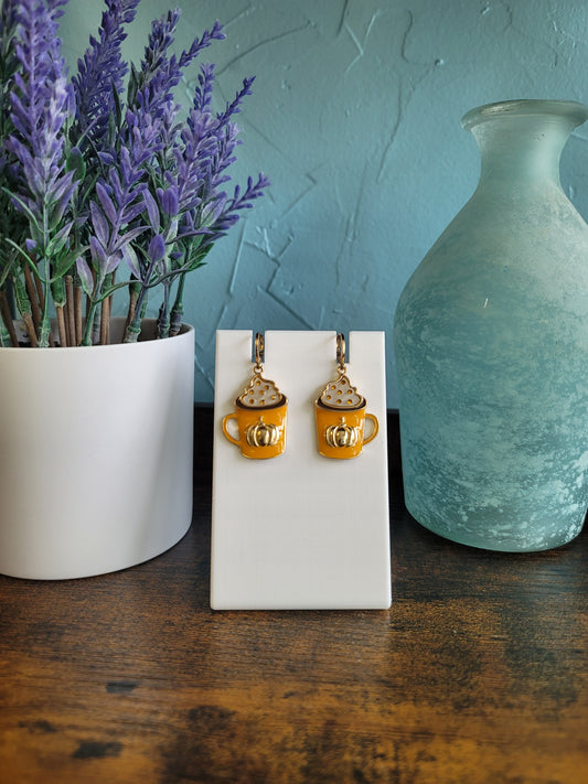 A pair of gold-colored earrings designed to resemble coffee beans, displayed on a white stand, with a lavender plant and a blue vase in the background.