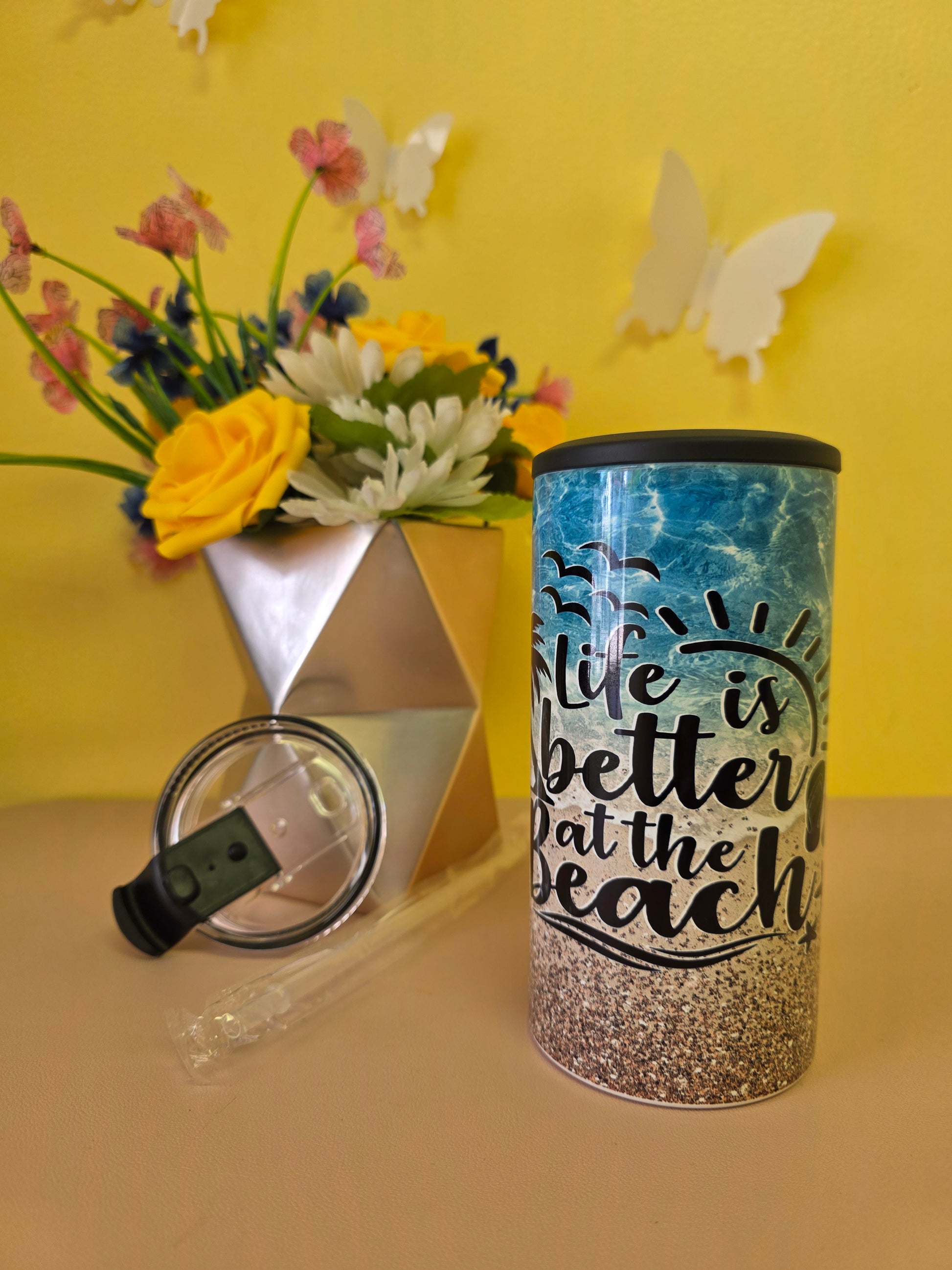 Insulated 4‑in‑1 stainless steel tumbler with sandy beach, ocean waves, and palm tree design, shown with lid, straw, and decorative flowers