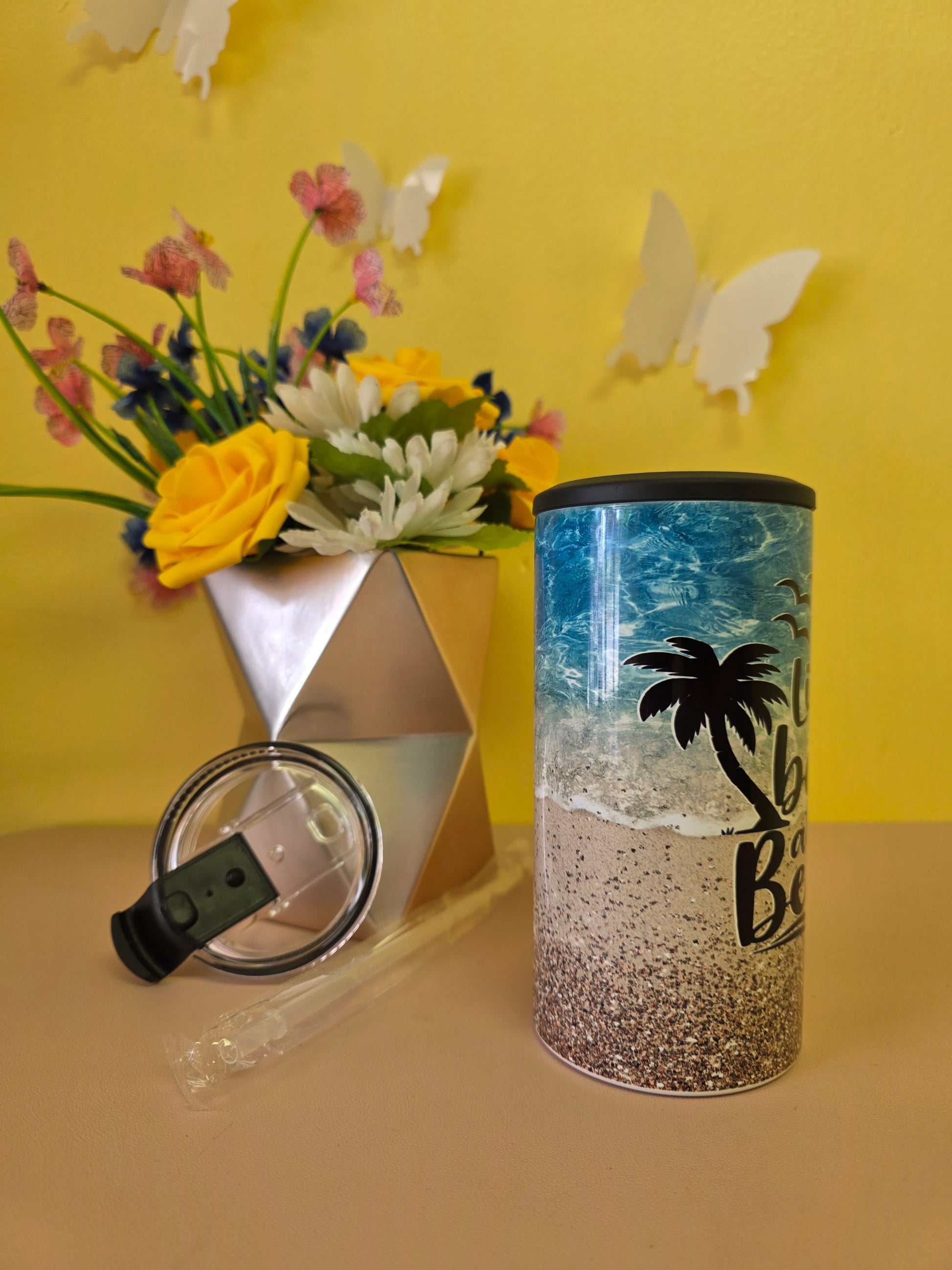Insulated 4‑in‑1 stainless steel tumbler with sandy beach, ocean waves, and palm tree design, shown with lid, straw, and decorative flowers