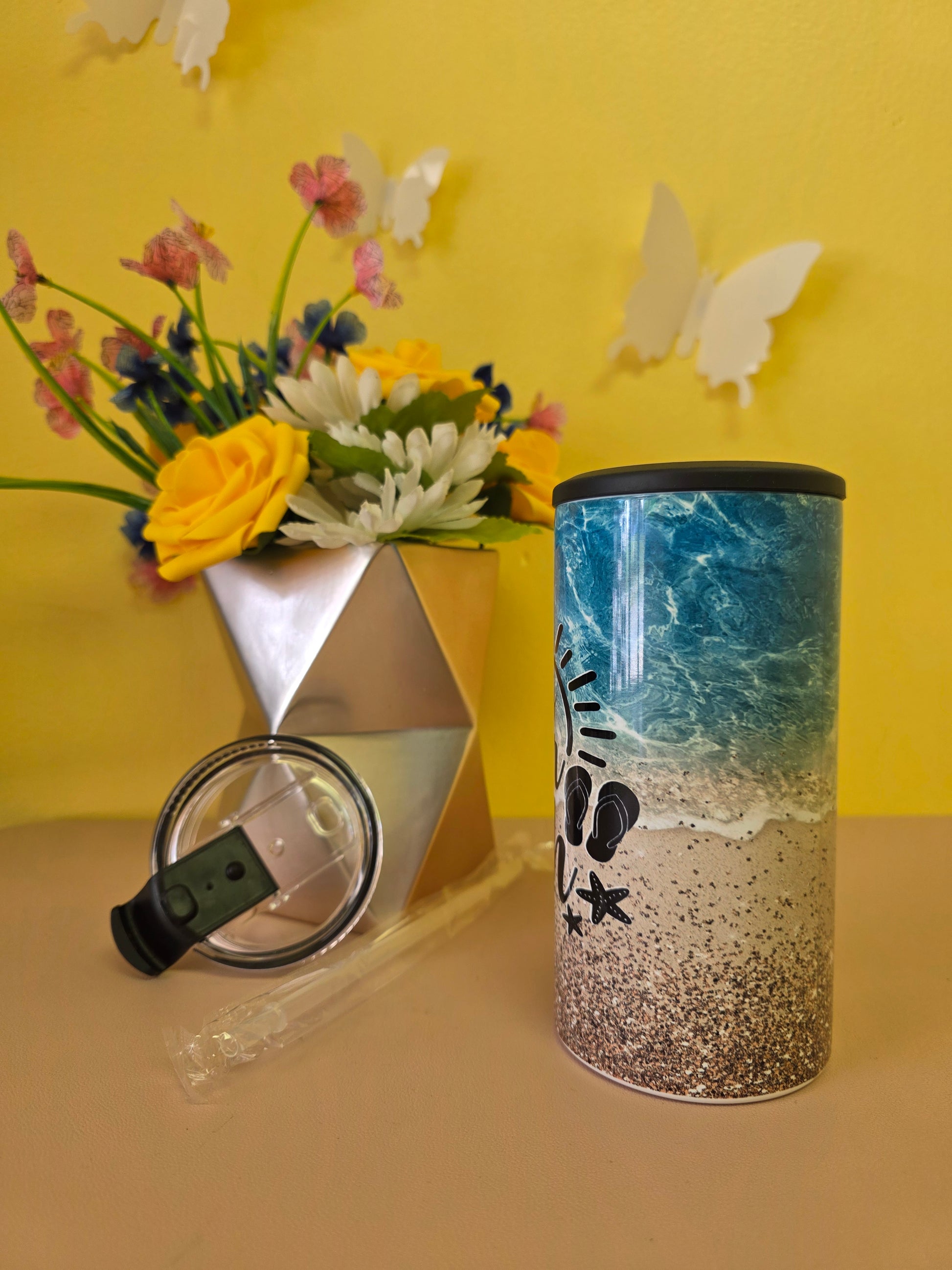 Insulated 4‑in‑1 stainless steel tumbler with sandy beach, ocean waves, and palm tree design, shown with lid, straw, and decorative flowers