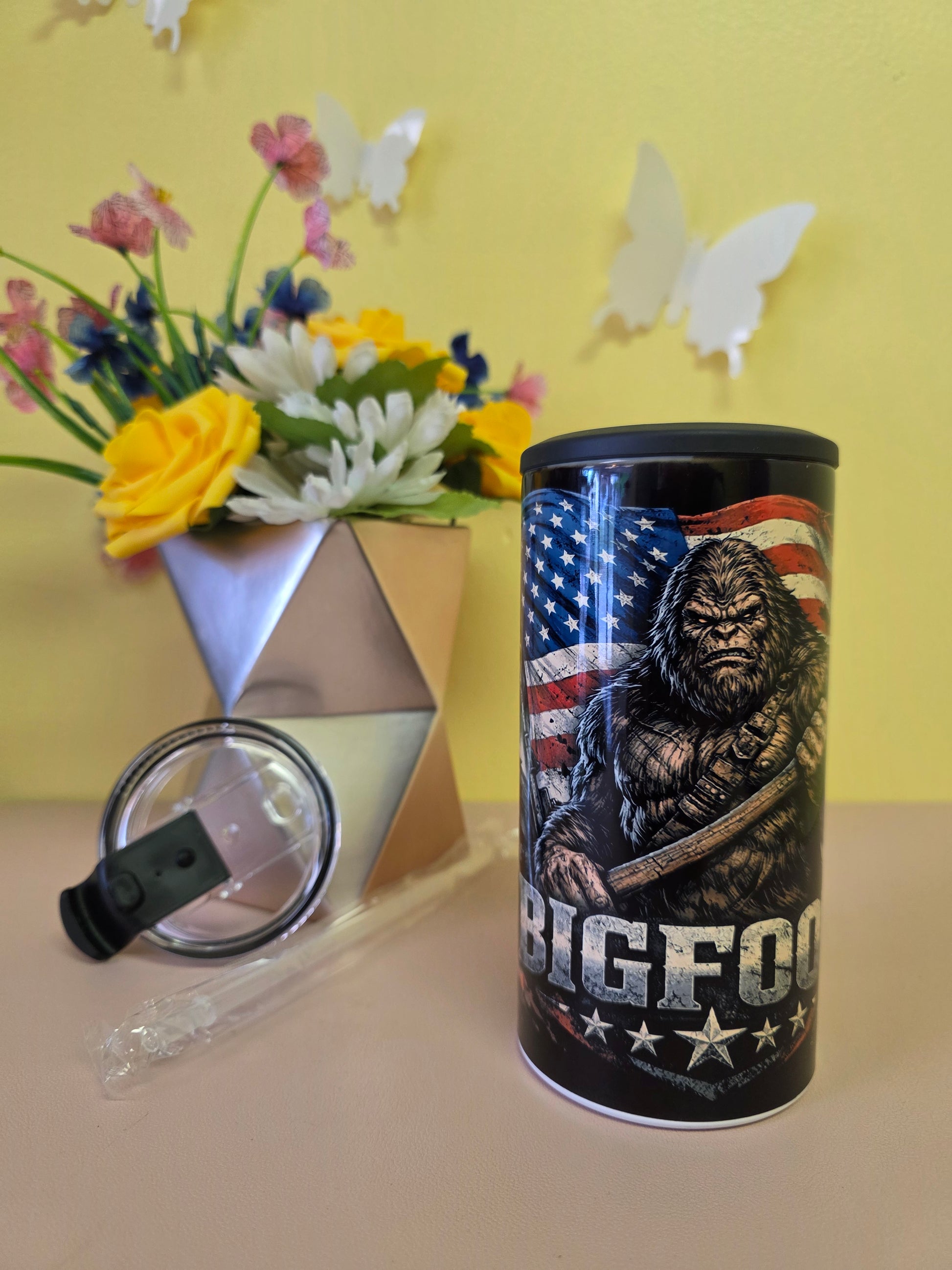 Black insulated stainless-steel tumbler with eagle and American flag artwork, shown with lid, straw, and floral background