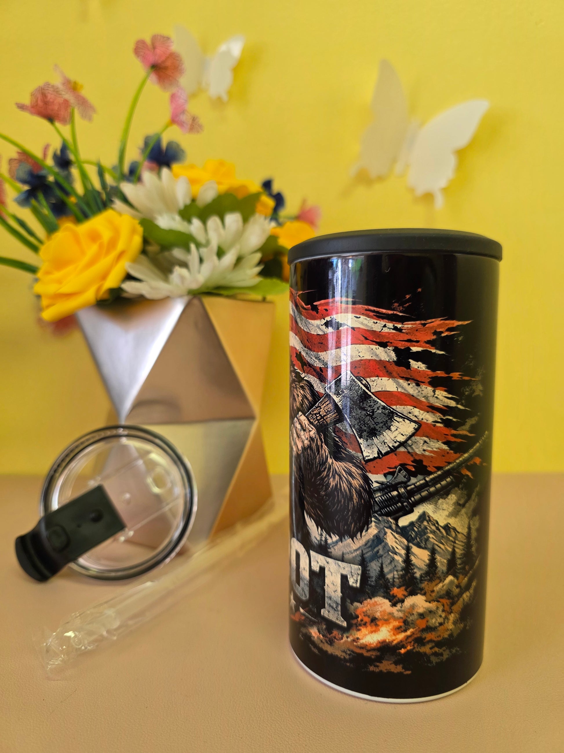 Black insulated stainless-steel tumbler with eagle and American flag artwork, shown with lid, straw, and floral background