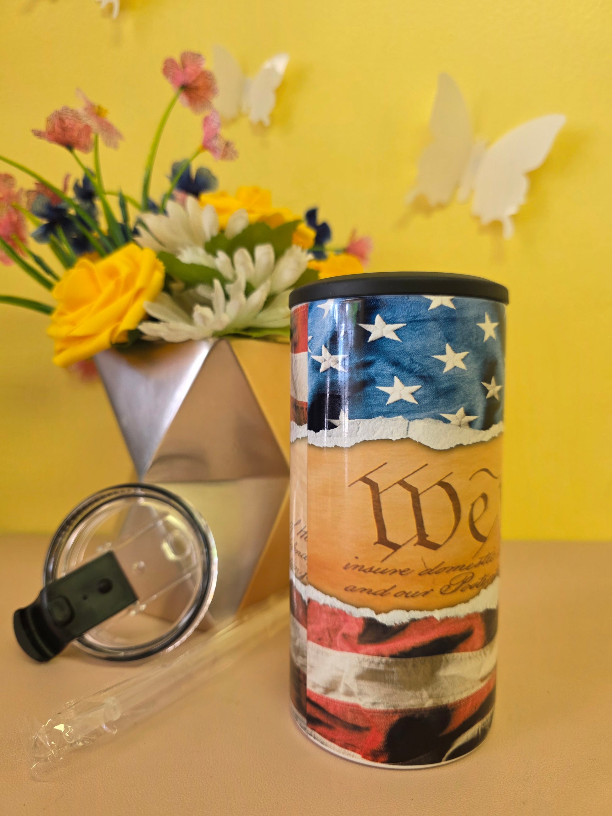 Insulated 4‑in‑1 stainless steel tumbler with U.S. Constitution text and American flag design, shown with lid, straw, and floral arrangement