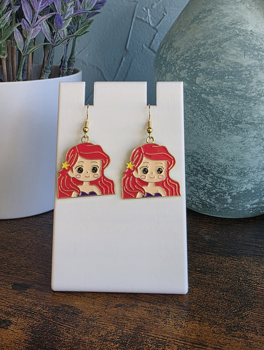 Cartoon-style earrings of a girl with red hair on a white stand with a plant and vase in the background.