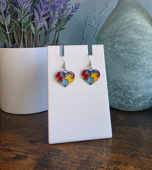 Heart-shaped earrings with colorful puzzle pieces for Autism on a white card, with a plant and vase in the background.