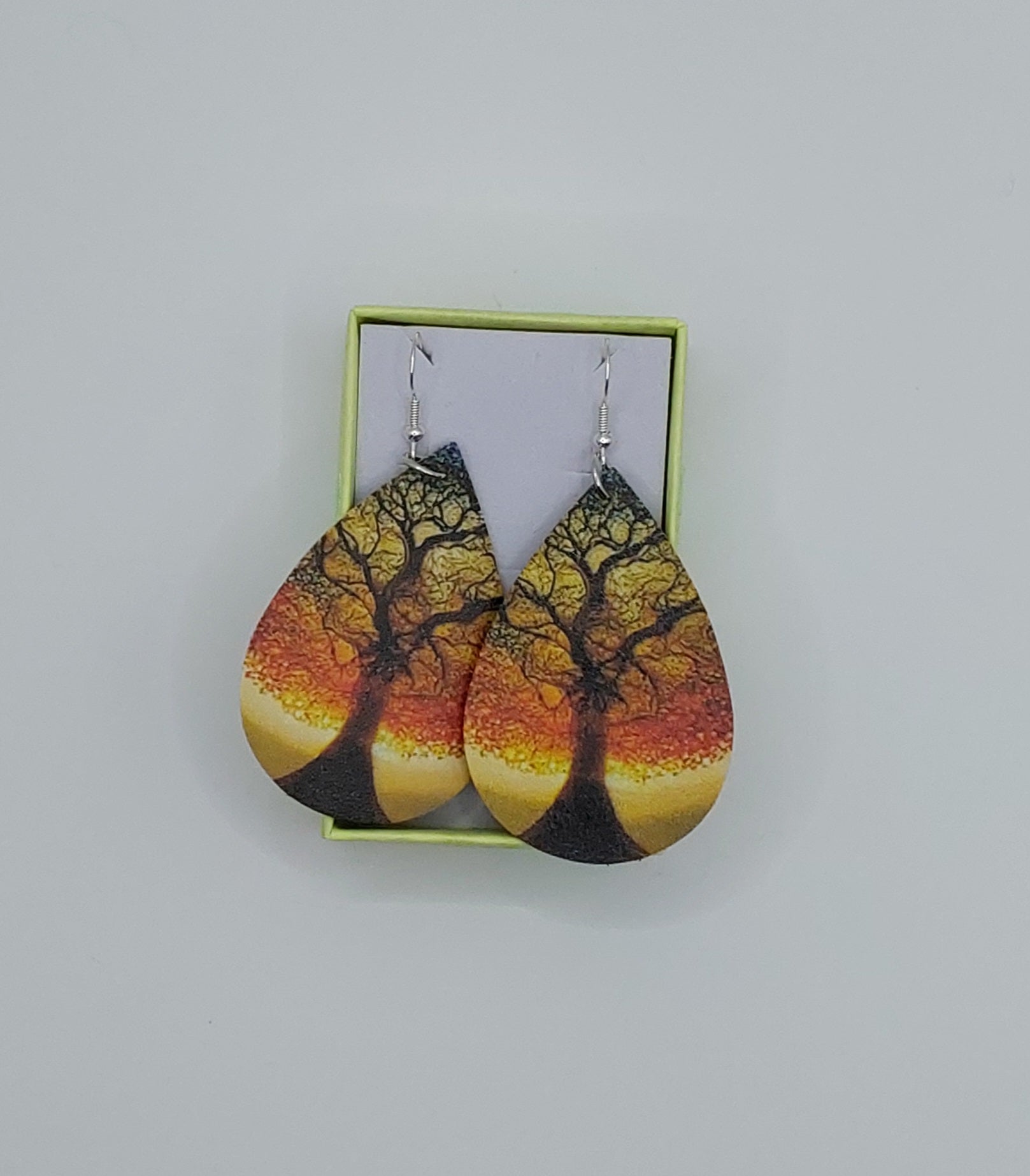 A pair of teardrop-shaped earrings with a autumnal tree pattern displayed in a small green box.