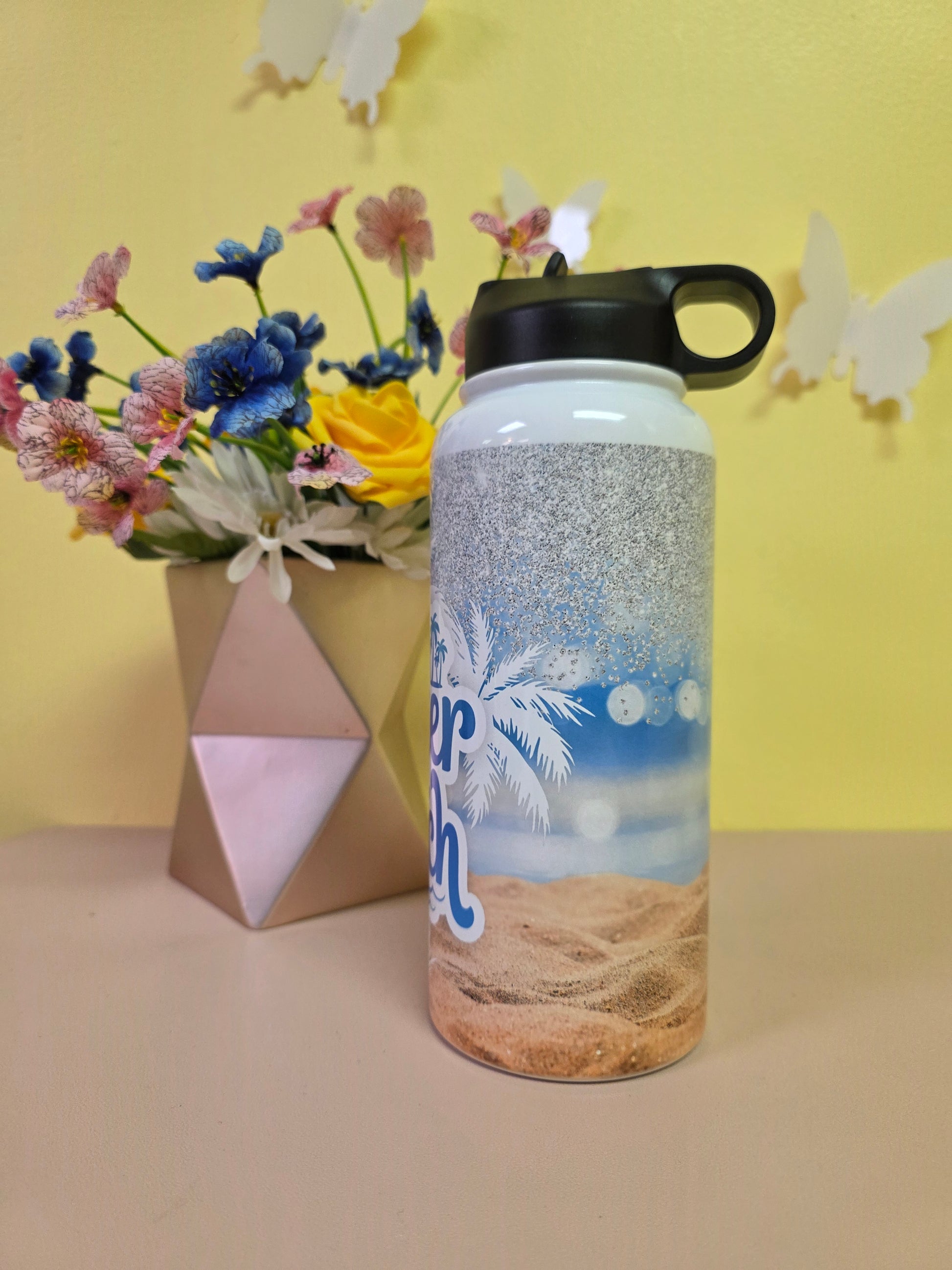 Water bottle with beach design on a yellow surface with flowers and a box.