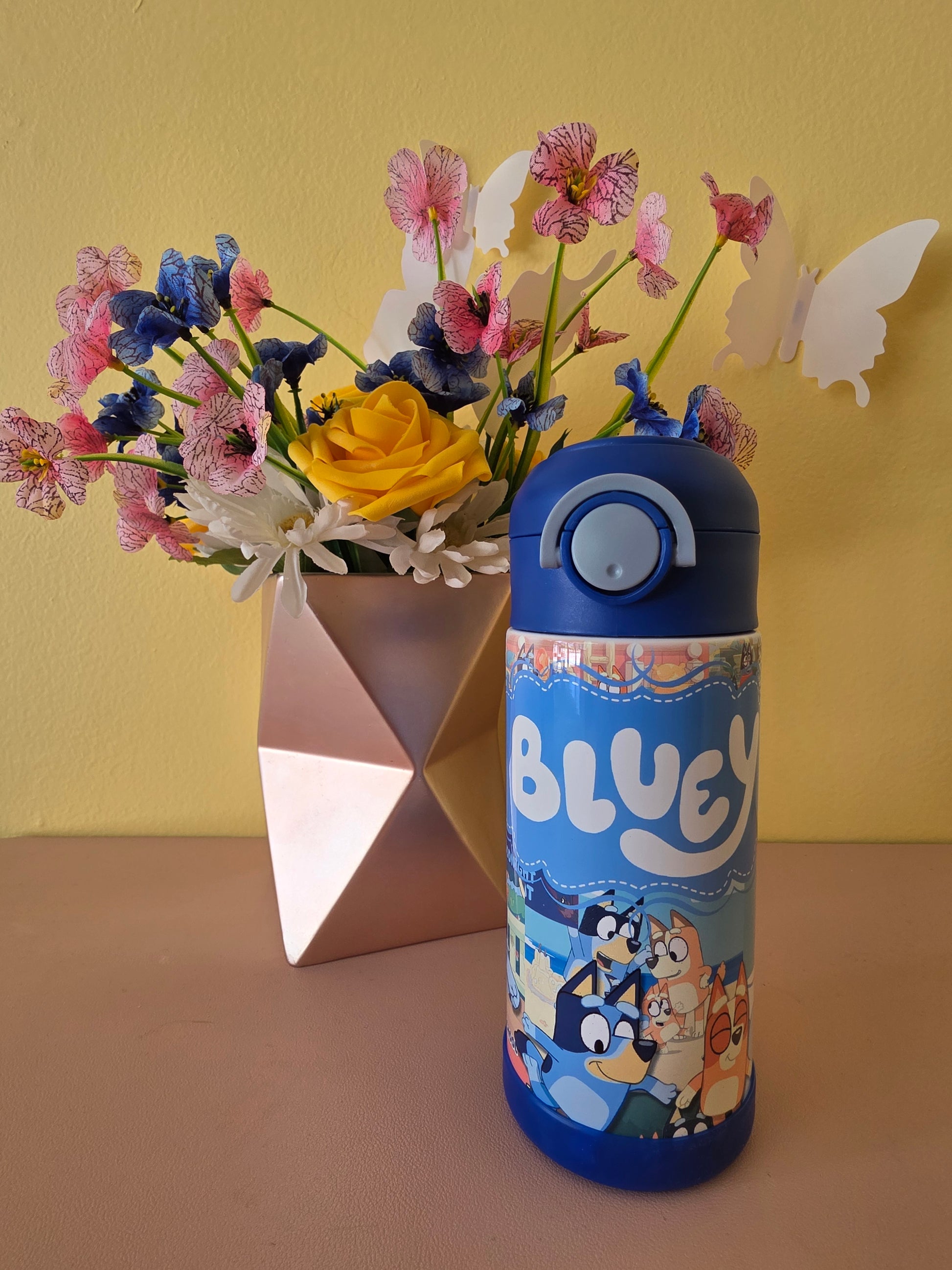 Bluey tumbler water bottle with lid and straw on a table with flowers in the background 