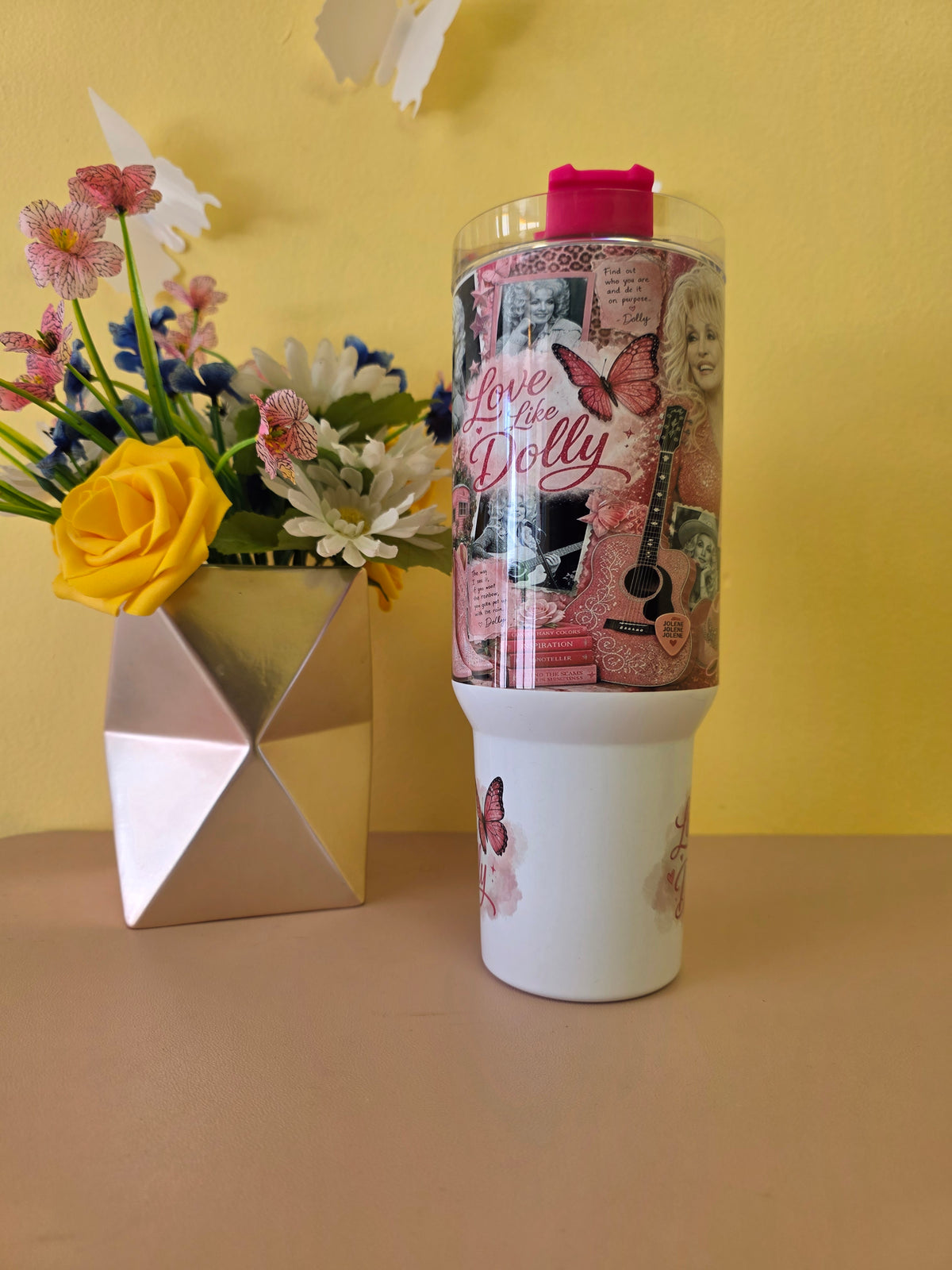 A white tumbler with a pink lid and a decorative collage wrap featuring 'Love for Dolly' text, butterflies, and musical instruments, set on a beige surface against a yellow wall next to a geometric vase of flowers.