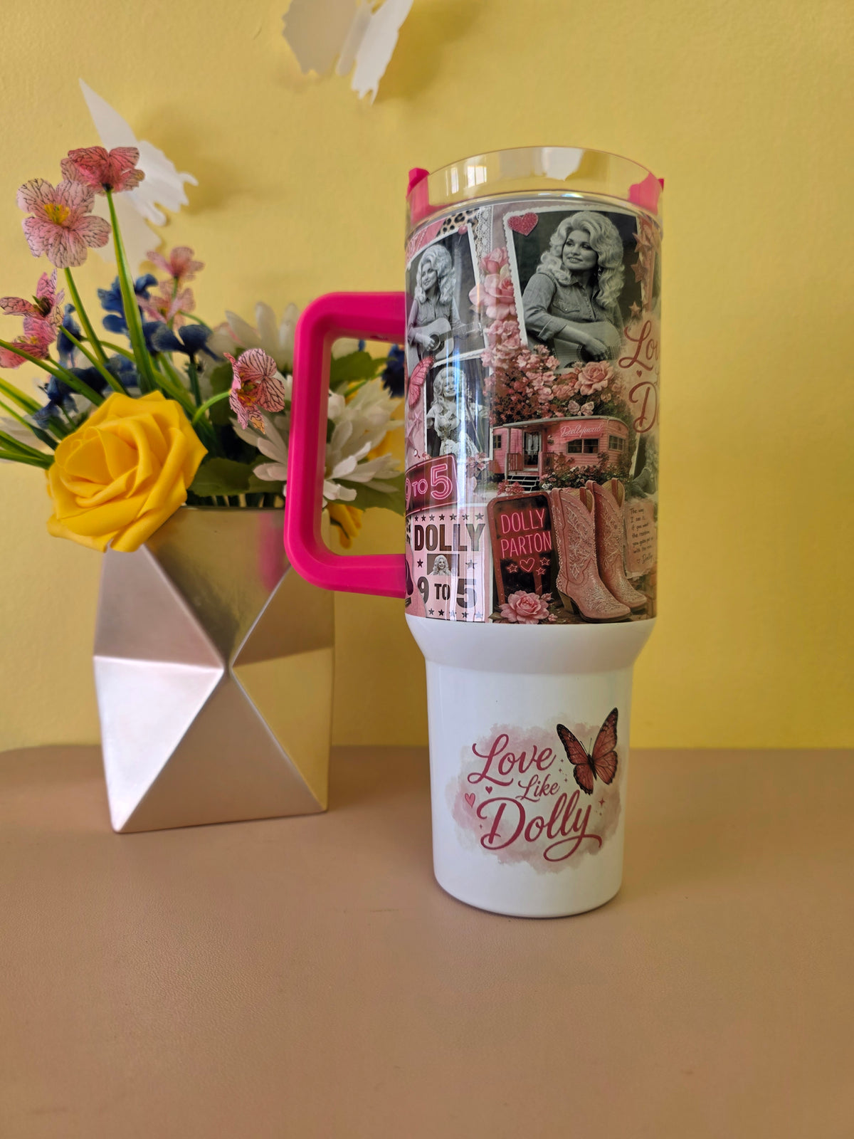 A white insulated tumbler with a bright pink handle and a decorative collage wrap featuring vintage Dolly Parton imagery, set on a beige tabletop against a yellow wall. A silver geometric vase with flowers sits to the left.