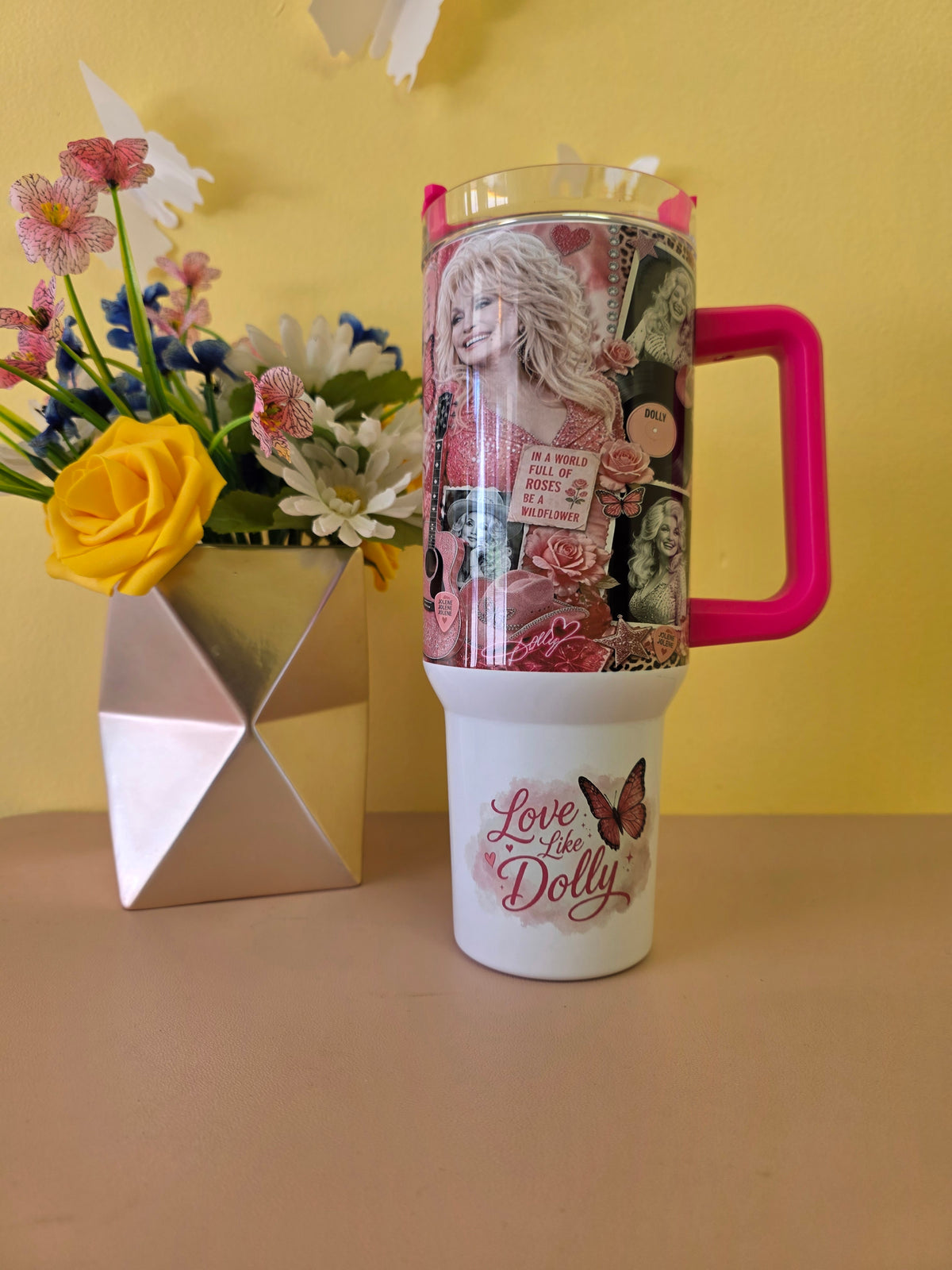 A tall insulated tumbler with a clear printed top section featuring a collage of vintage-style portraits and pink floral text, attached to a white lower section with the words 'Love the Dolly' and a butterfly. The tumbler has a bright pink handle and sits on a beige tabletop against a yellow wall, next to a metallic vase with flowers.