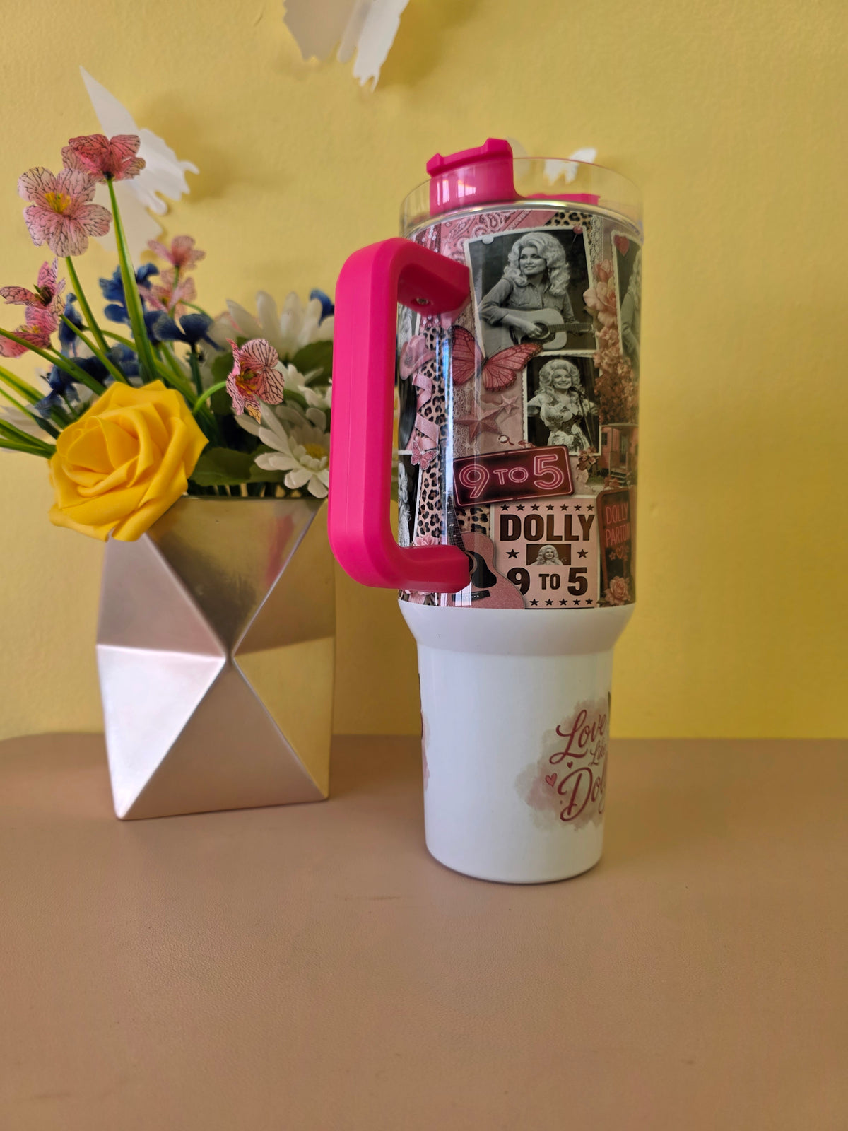 A white insulated tumbler with a bright pink handle and lid, decorated with a collage of vintage-style portraits and text, standing on a beige tabletop against a yellow wall. A metallic vase with flowers sits to the left.