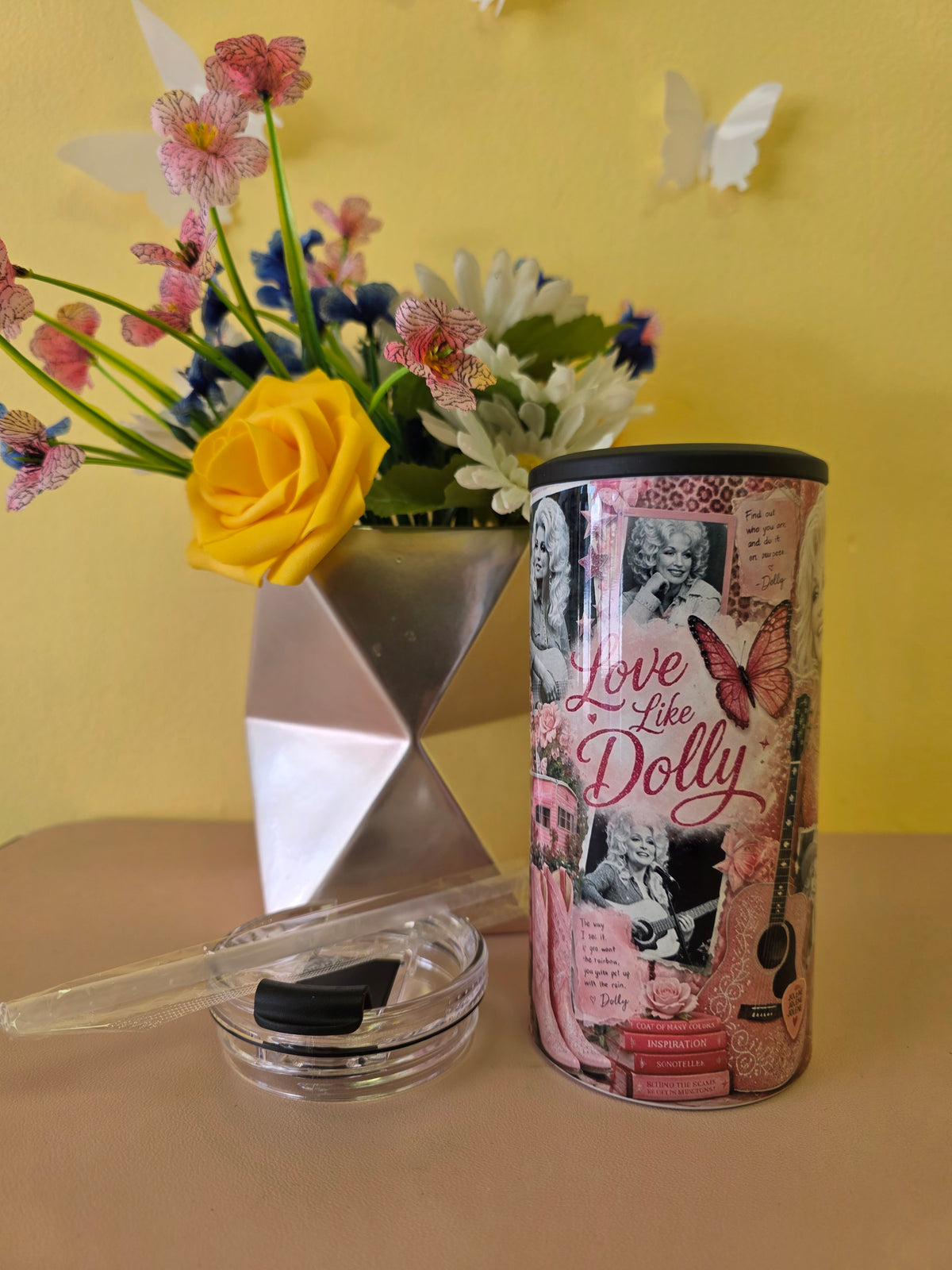 A cylindrical cosmetic container with a pink collage design reading 'Love like Dolly' sits on a beige surface next to a small clear dish with a black lid. A metallic vase with flowers is blurred in the background against a yellow wall.
