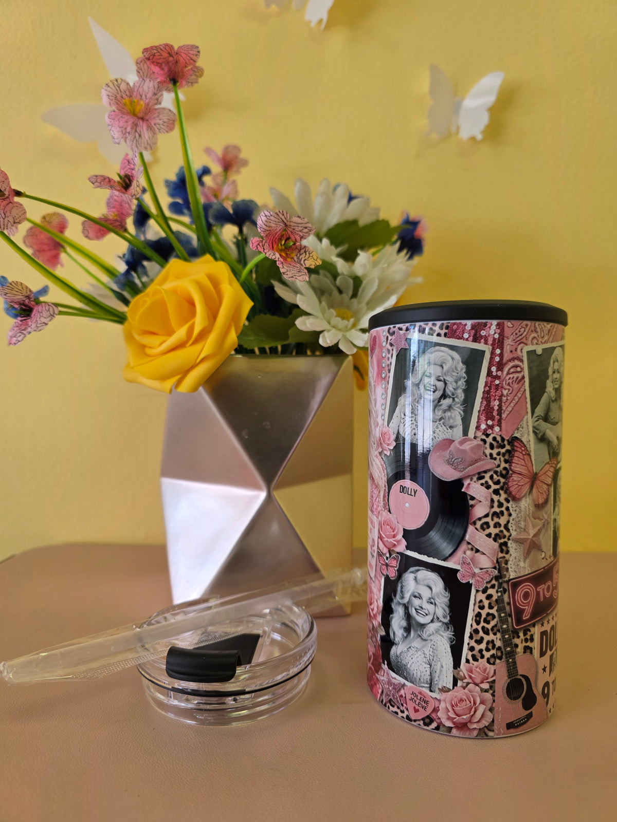 A pink and black decorative cylindrical tumbler with a collage of vintage-style portraits, roses, butterflies, and vinyl records, shown next to a small clear glass jar with a black lid and a clear straw. The scene is styled with a geometric silver vase of flowers against a yellow wall.