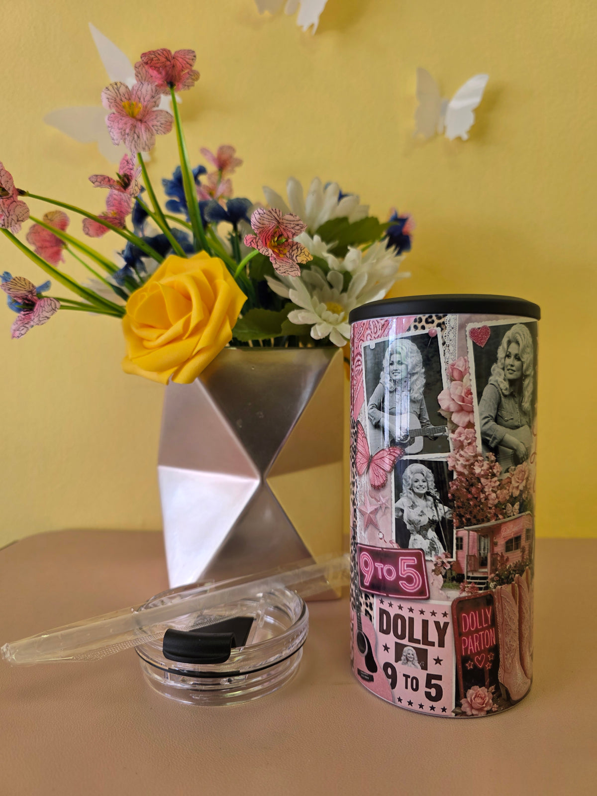 A cylindrical cosmetic container with a black lid and a pink collage-style label featuring Dolly Parton imagery, placed on a beige surface next to a small clear jar with a black lid and a clear applicator. A geometric silver vase with flowers sits against a yellow wall.
