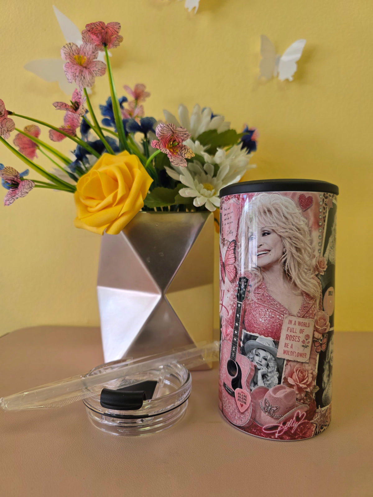 A cylindrical tin of Dolly Parton memorabilia sits on a beige tabletop next to a small clear glass dish with a black lid and a clear plastic tool. The tin features a collage of photos, guitars, roses, and text, with a picture of Dolly Parton prominently displayed.
