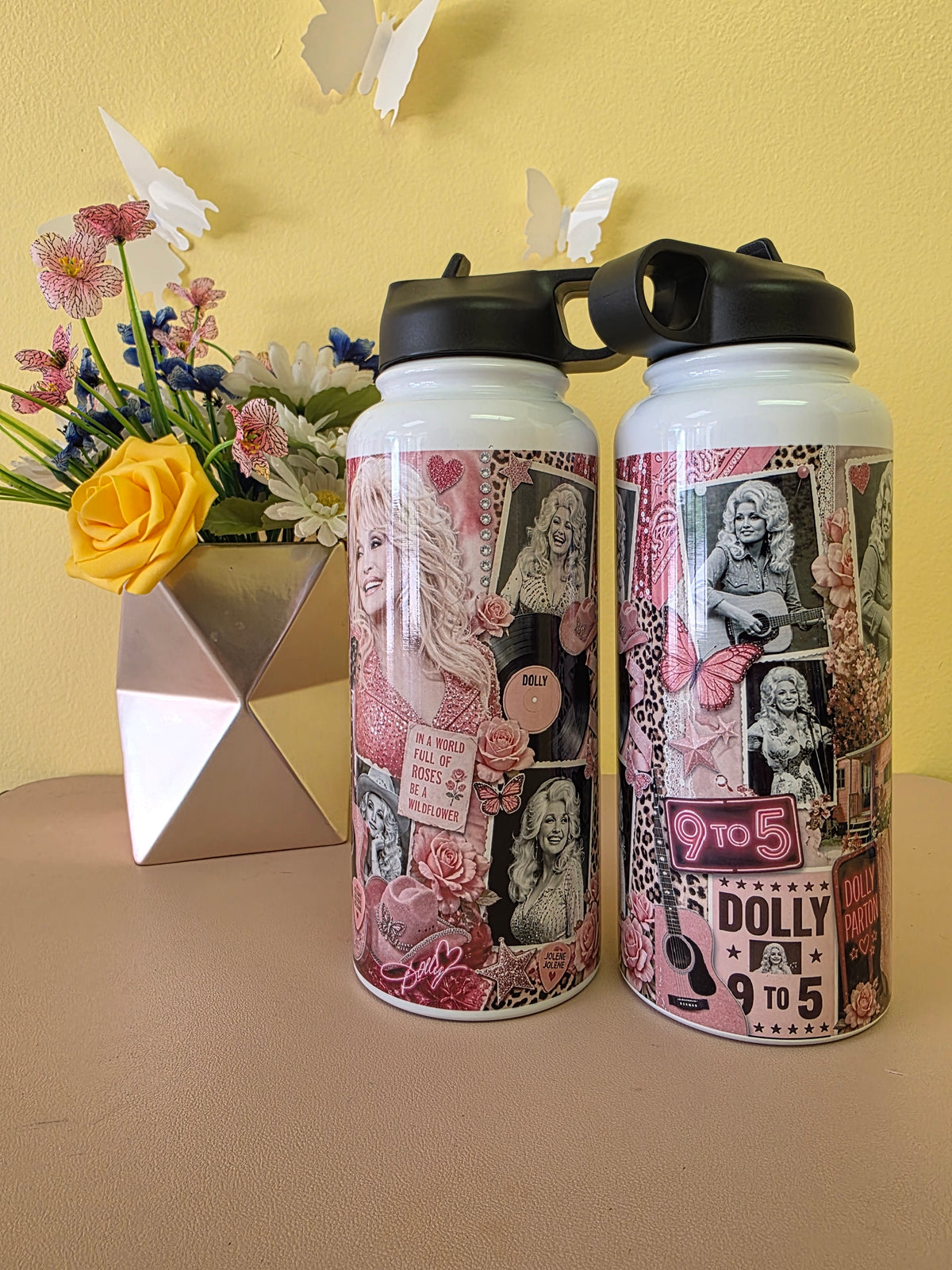 Two white insulated water bottles with black lids, decorated with pink and black collage graphics featuring portraits and text, displayed on a beige surface against a yellow wall with a floral vase in the background.