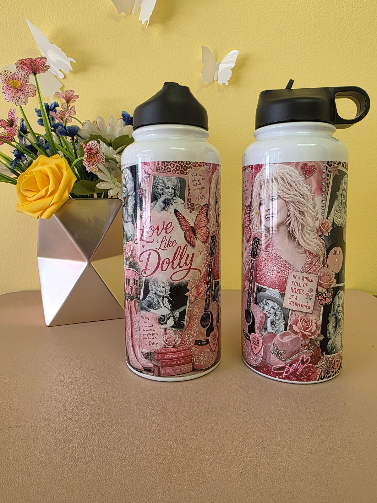 Two white insulated water bottles with black lids, decorated with pink and black collage artwork featuring the text 'Love like Dolly' and floral/butterfly motifs, displayed on a beige surface against a yellow wall with a flower vase in the background.