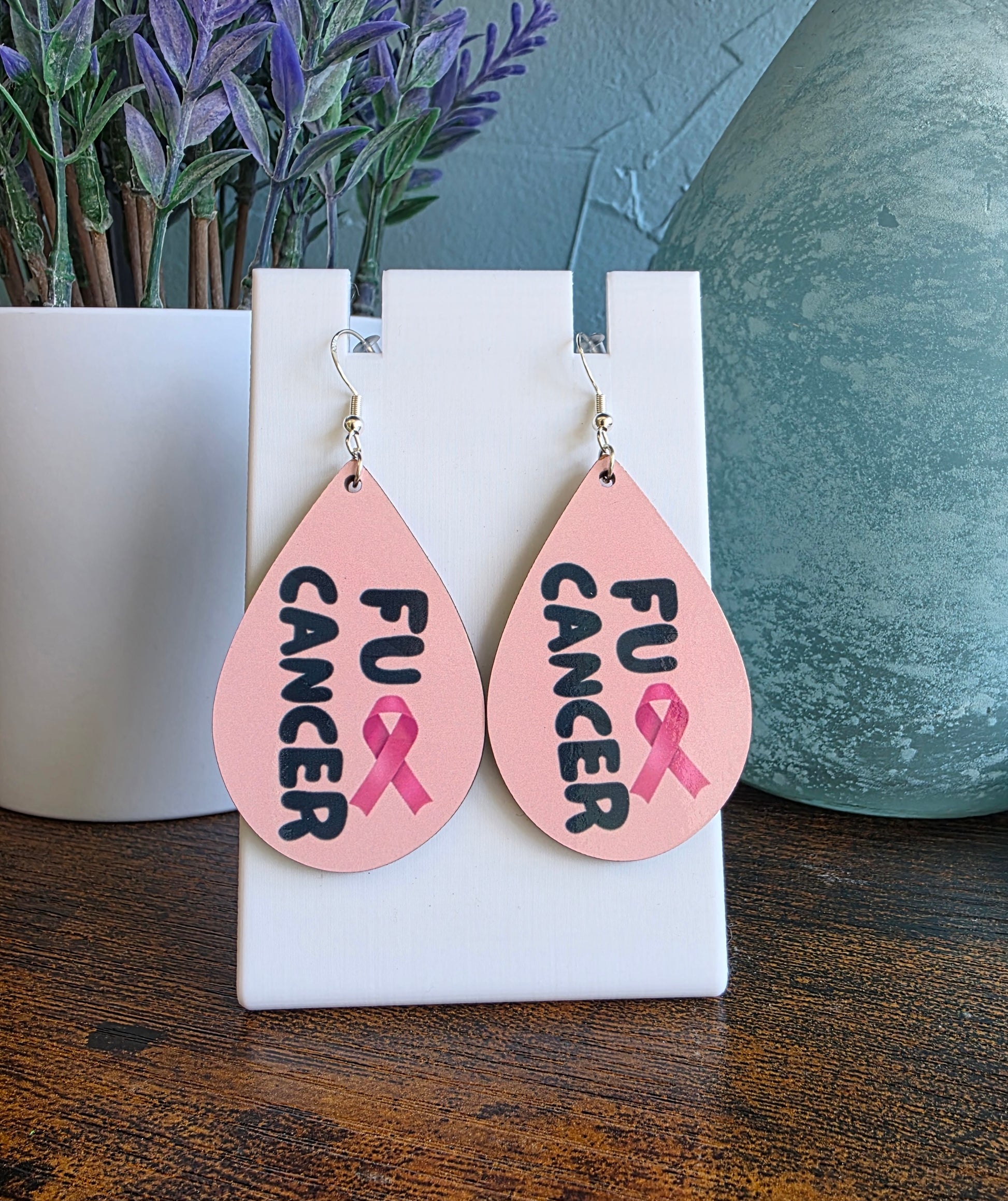 Teardrop-shaped earrings with 'FU CANCER' and pink ribbon design on a white stand.