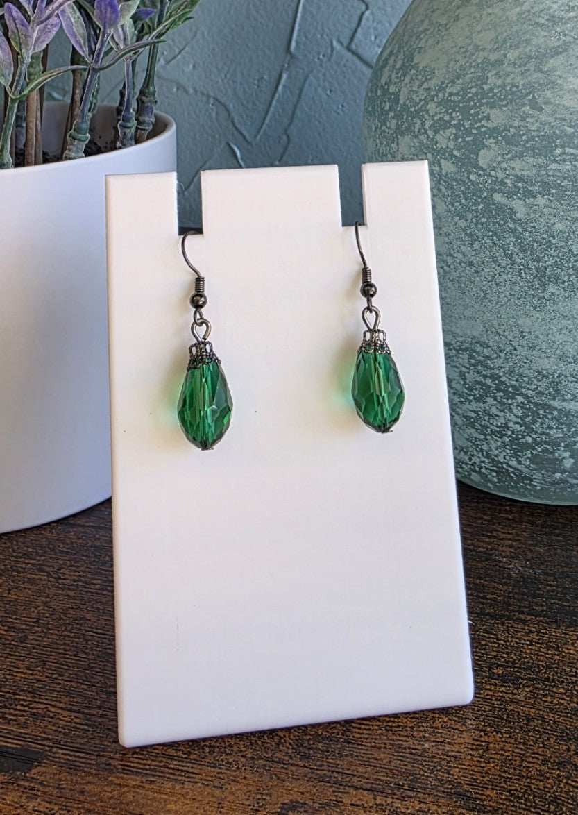 Green teardrop earrings on a white card with a plant and decorative object in the background.