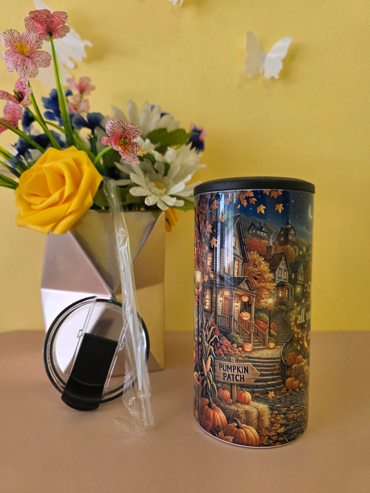 A cylindrical metal tin with a lid, featuring an autumn-themed illustration of a haunted house and pumpkins labeled 'Pumpkin Patch,' displayed next to a vase of flowers and a clear straw on a beige surface against a yellow wall.
