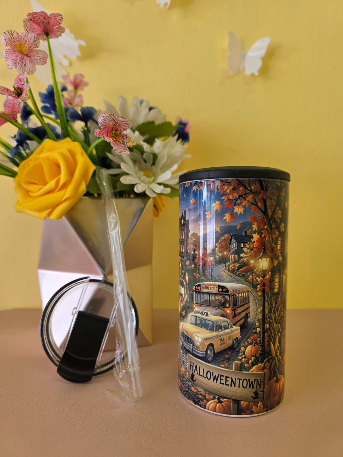 A cylindrical tumbler with a Halloween-themed printed design featuring a yellow school bus, taxi, pumpkins, and autumn leaves, standing next to its clear lid on a beige surface against a yellow wall. A vase of flowers is blurred in the background.