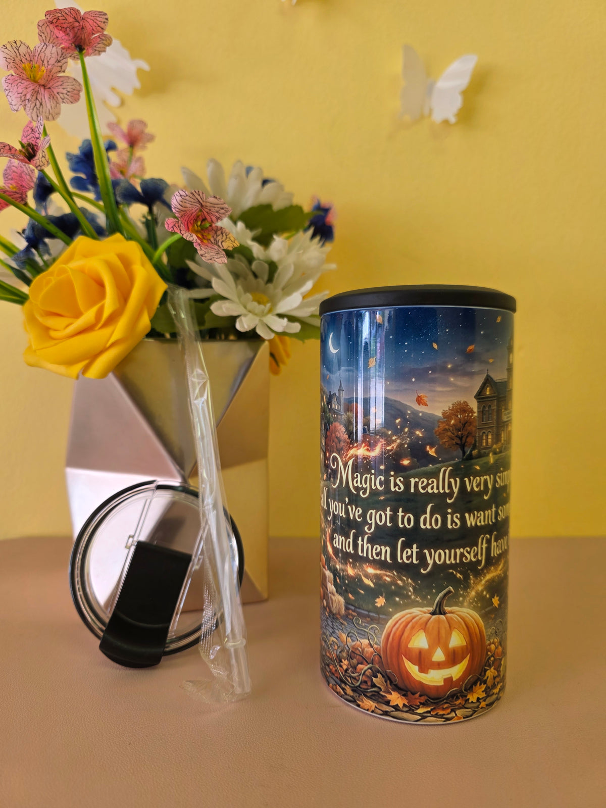 A cylindrical tumbler with a Halloween-themed printed design featuring a jack-o'-lantern and autumn scene, shown next to its clear lid and straw on a beige surface against a yellow wall. A vase of flowers is blurred in the background.