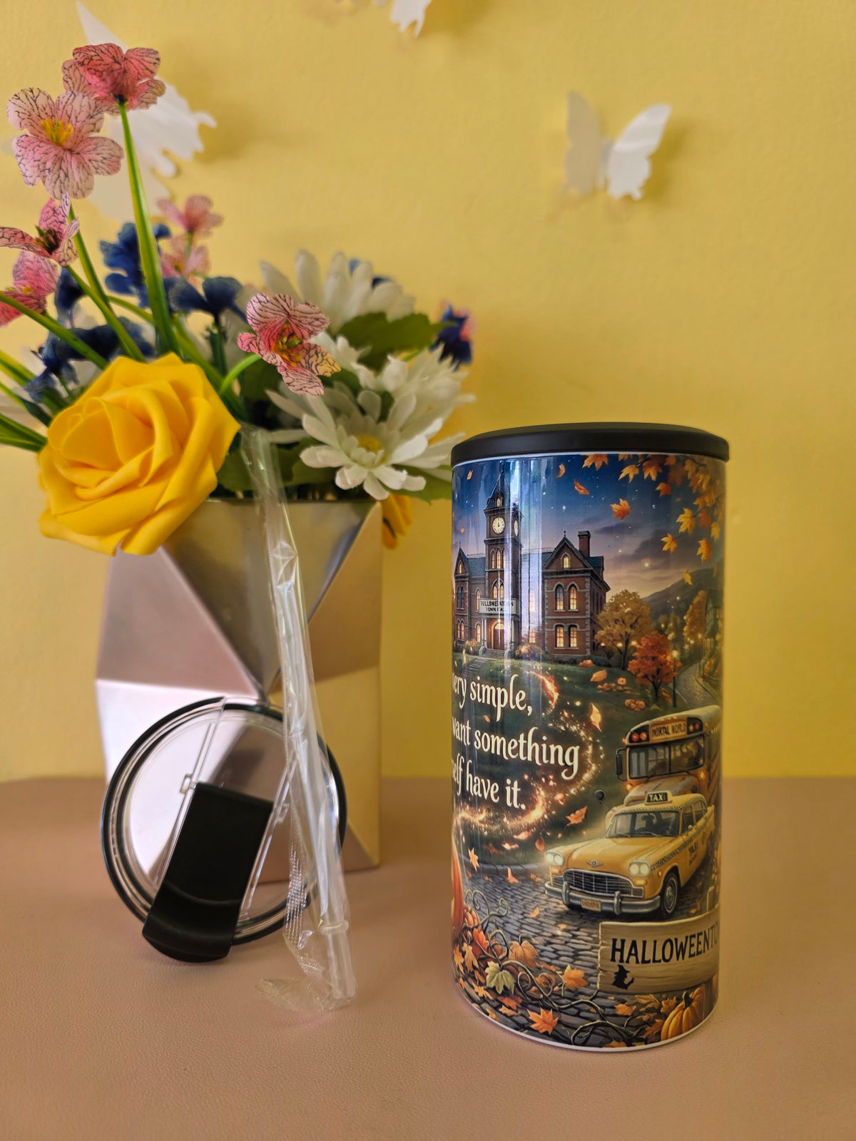 A cylindrical metal canister with a Halloween-themed printed label stands on a tabletop next to its black lid. The label features a haunted house, a yellow taxi, and autumn leaves, with partial text including 'Halloween' and 'stay simple, stay something'.