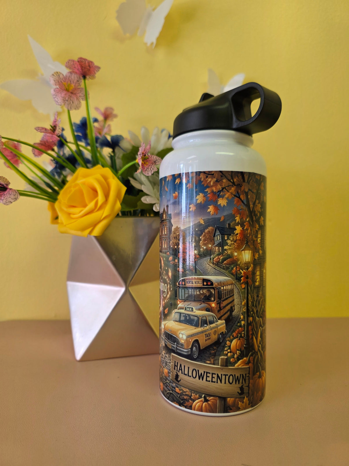 A white insulated water bottle with a black lid and a detailed autumn Halloween illustration featuring a yellow taxi, pumpkins, and the text 'Halloween Town' and 'Welcome'. It is placed on a beige surface next to a geometric silver vase with flowers against a yellow wall.