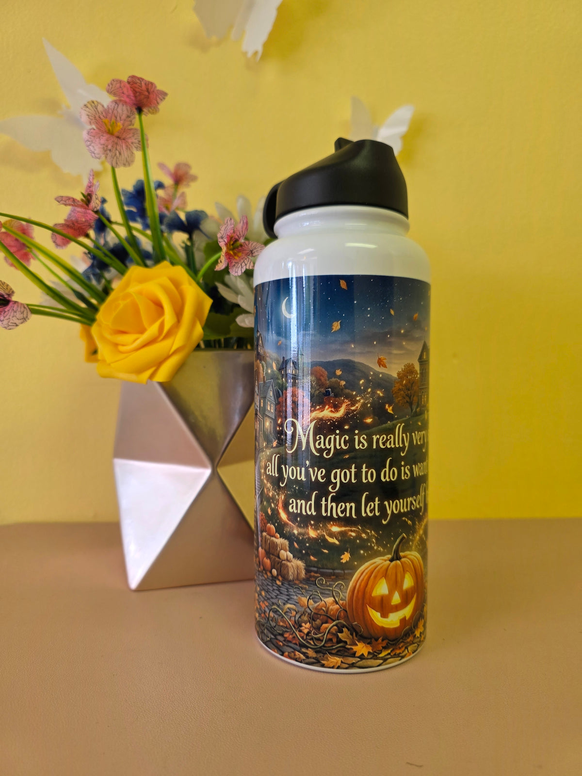 A white insulated water bottle with a black lid stands on a beige surface against a yellow wall. The bottle has a printed autumn scene with a glowing jack-o'-lantern and cursive text.