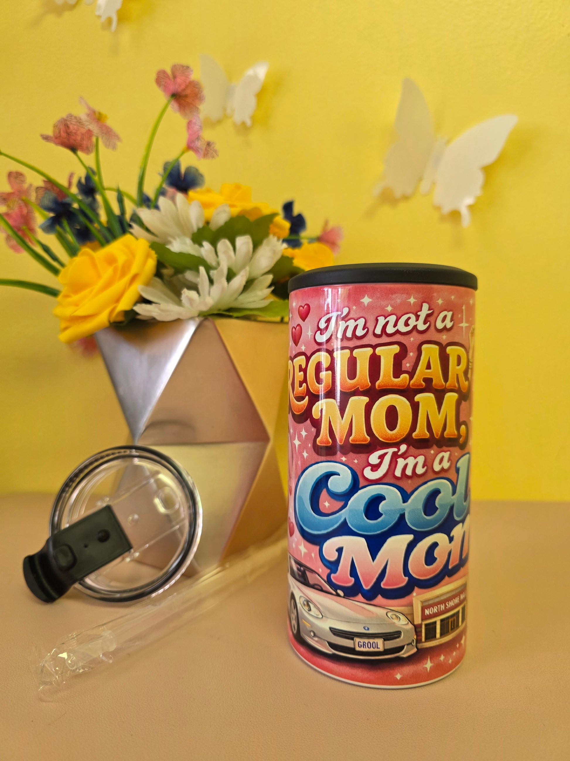 Pink insulated stainless steel tumbler with ‘I’m not a regular mom I’m a cool mom’ design, clear lid, straw, and floral background decor