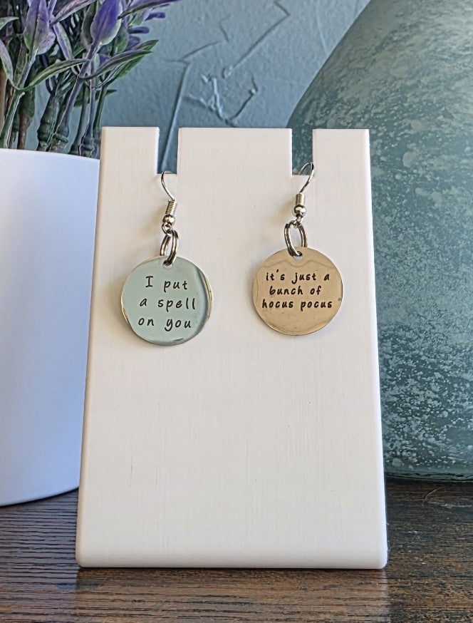 Pair of earrings with Hocus Pocus phrases on a white card against a neutral background