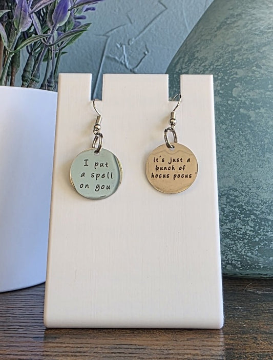 Pair of earrings with Hocus Pocus phrases on a white card against a neutral background
