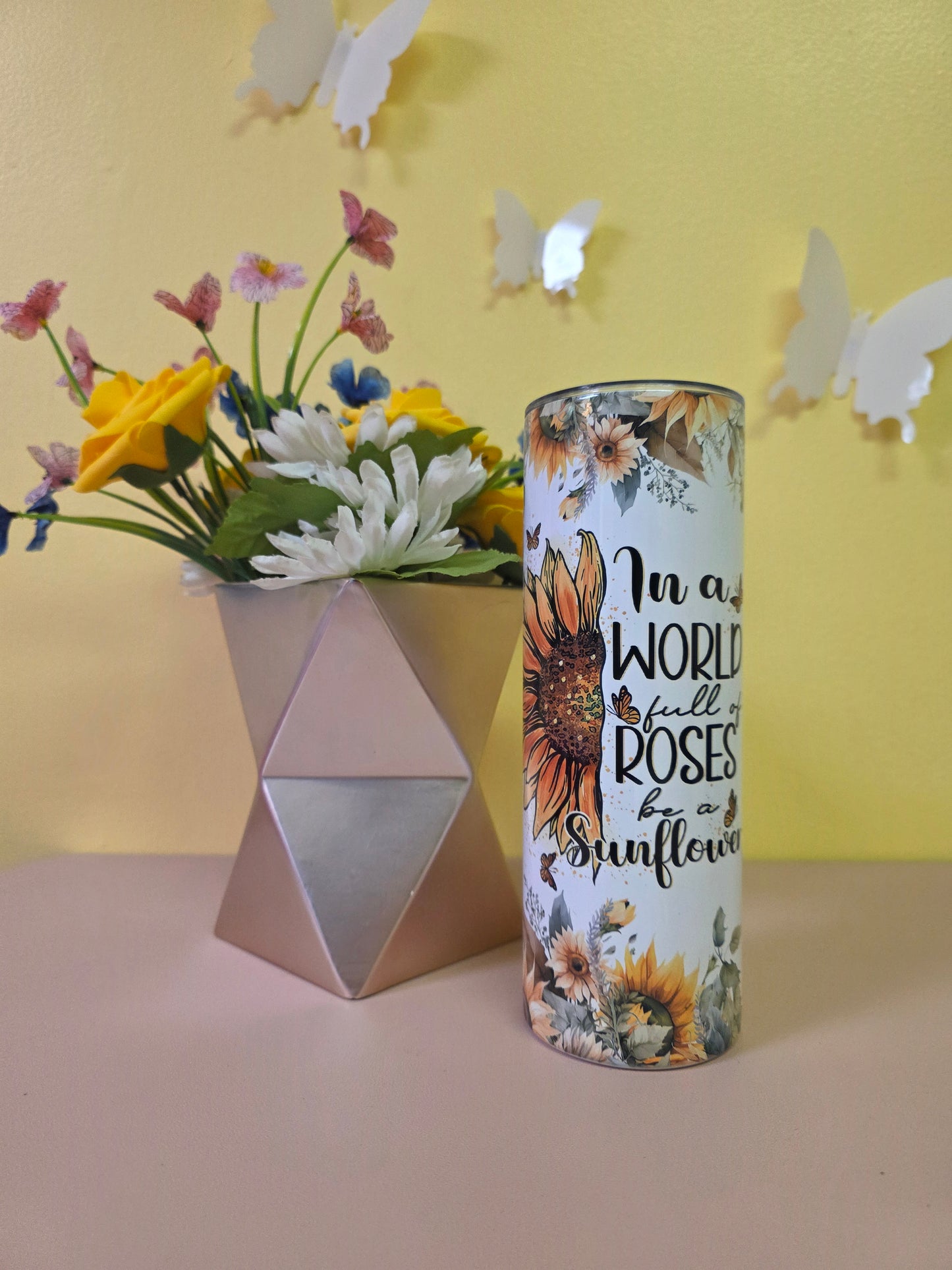 Decorative cylindrical container with floral design and text, placed on a yellow surface with flowers and a pink triangle.