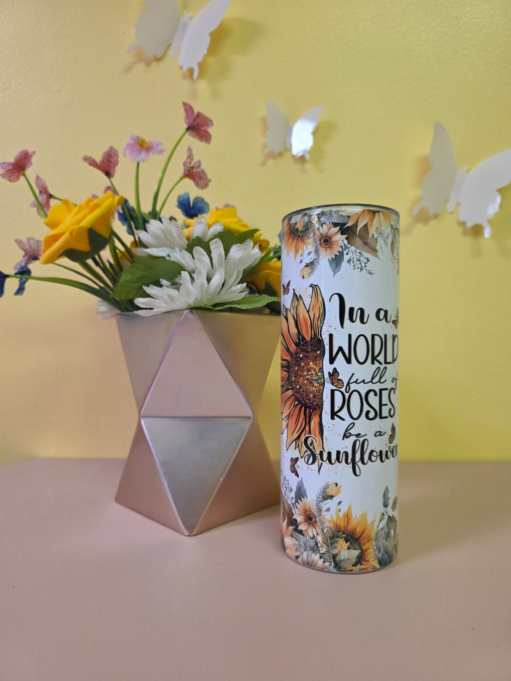 Decorative cylindrical container with floral design and text, placed on a yellow surface with flowers and a pink triangle.